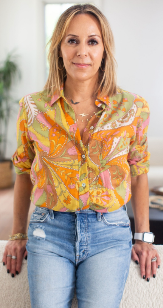 Kim Shapira standing indoors, wearing a colorful, patterned blouse and ripped jeans, with her hands resting on a surface behind her.