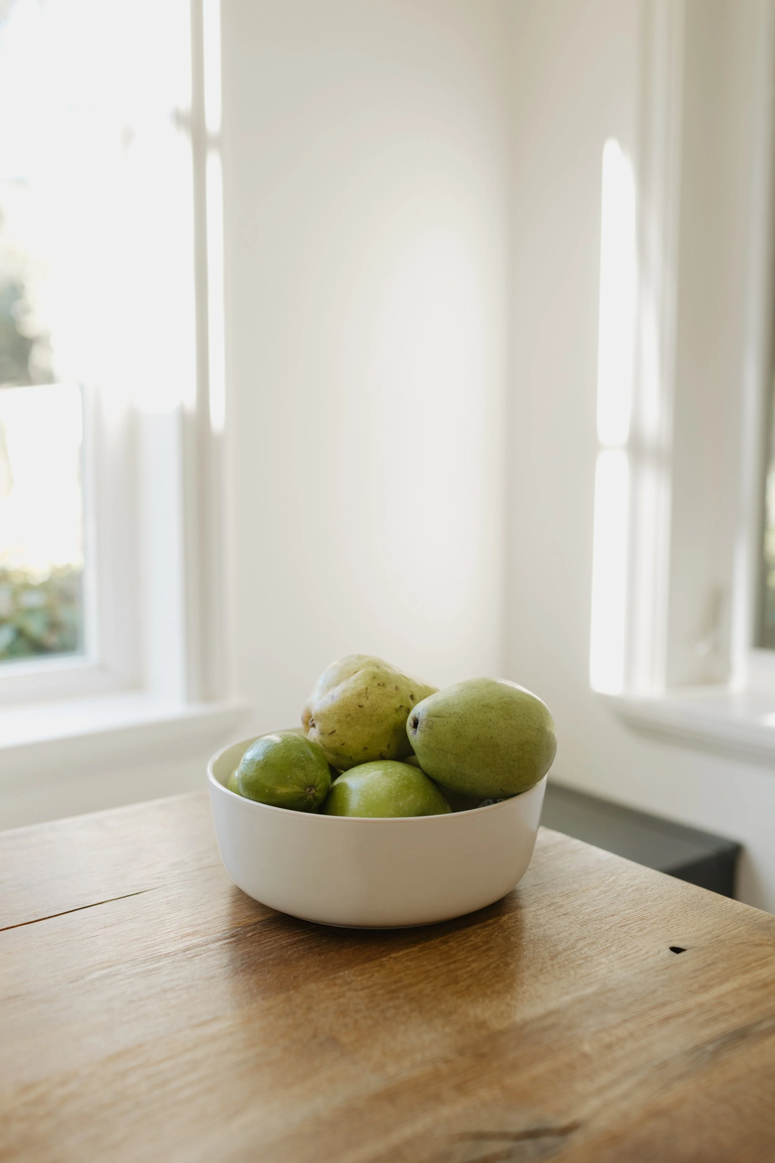 A white bowl filled with green apples and pears on a wooden table in a bright room with white walls and windows.