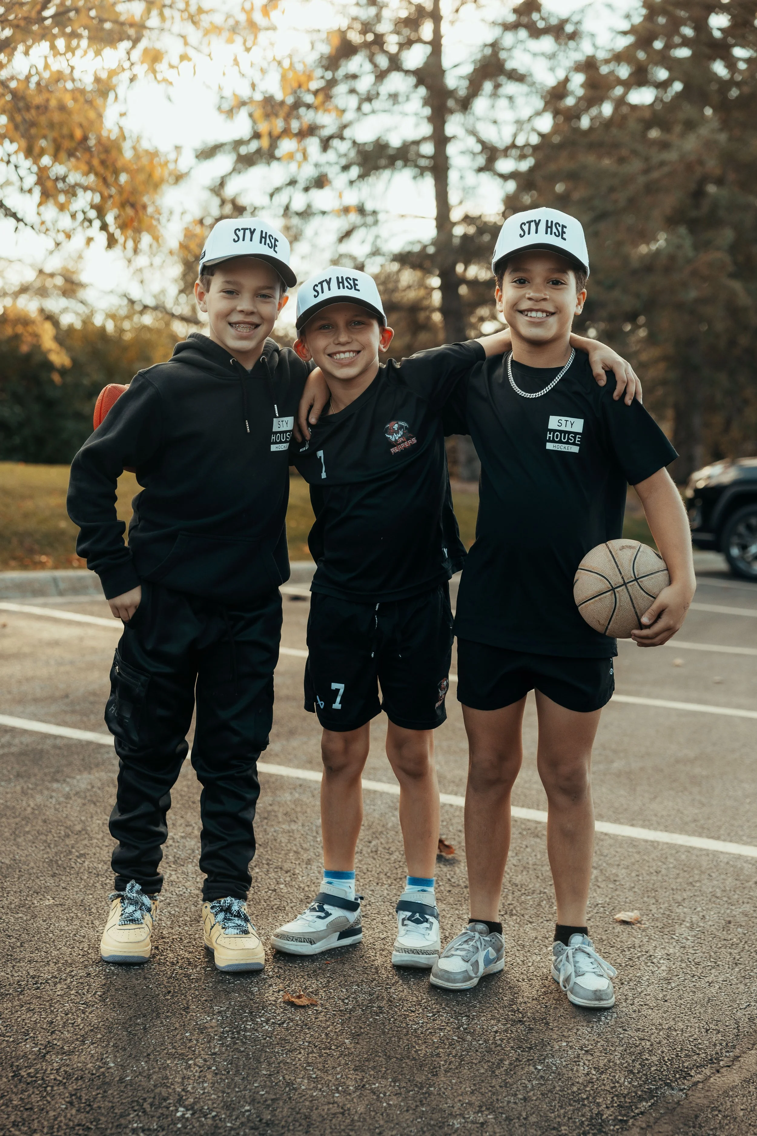 Three young boys standing on a basketball court outdoors, smiling and arm-in-arm, wearing matching white hats with black text, casual sportswear, and sneakers. One boy holds a basketball. There are trees with autumn leaves in the background.