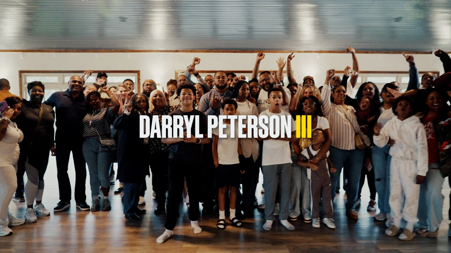 A large group of diverse people gathered indoors, smiling and cheering, with the text 'Darryl Peterson III' overlaid in the center.