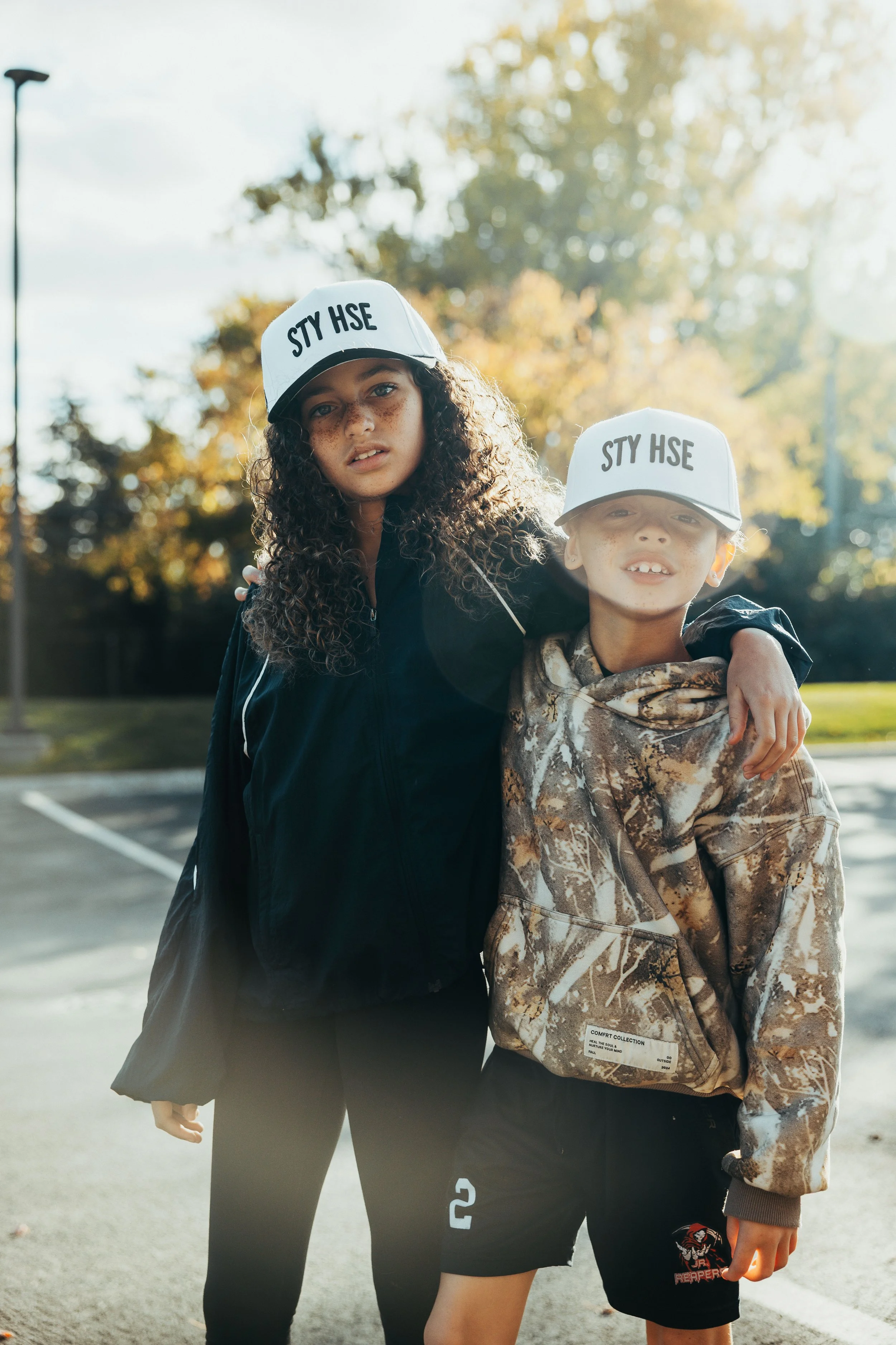 Two kids standing outdoors in a parking lot with trees in the background, both wearing white baseball caps that say "STY HSE," with one kid's arm around the other's shoulder, during daytime.