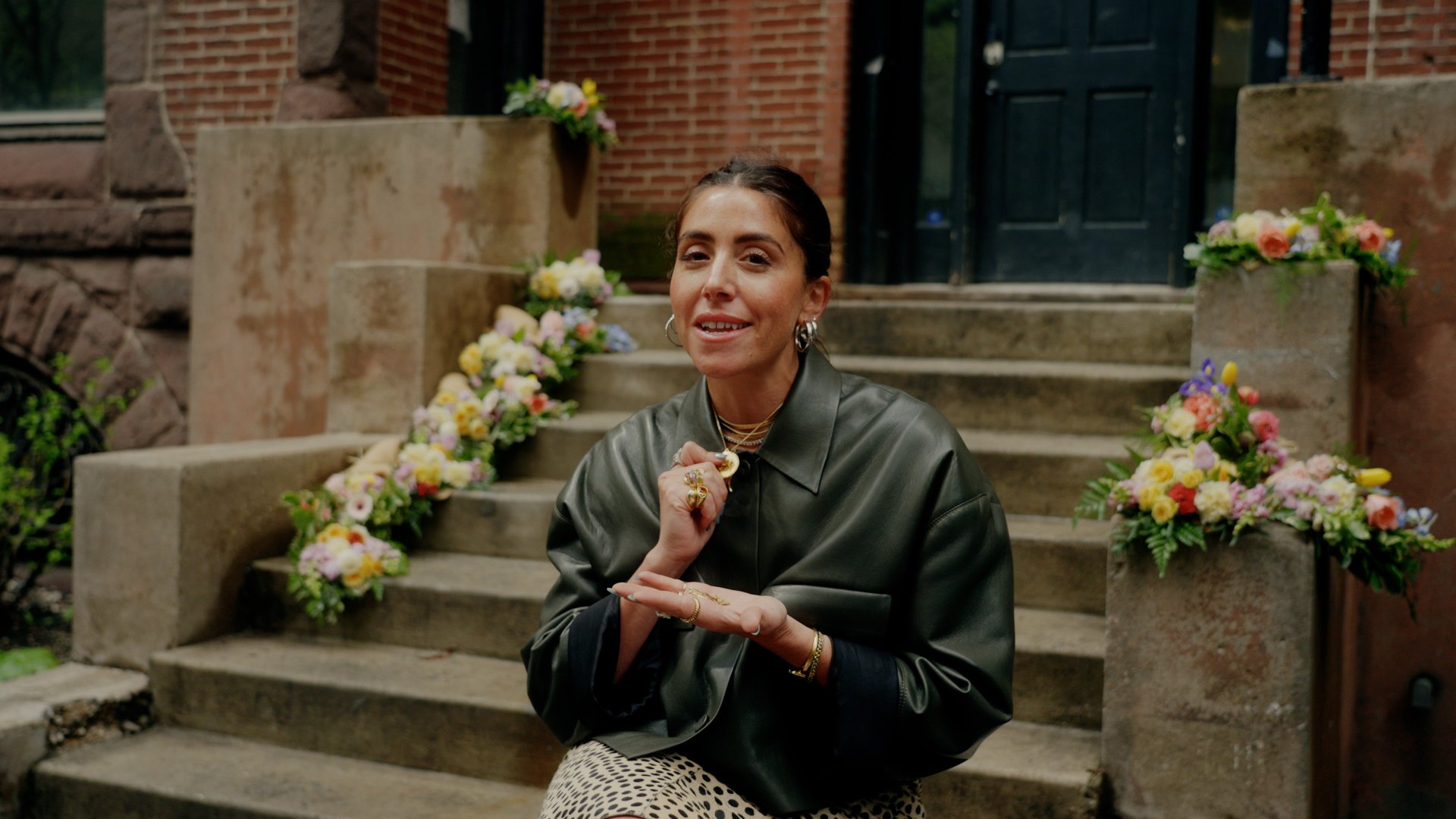 A woman with dark hair and hoop earrings sitting on stone steps outside, surrounded by colorful floral arrangements, wearing a dark leather jacket.