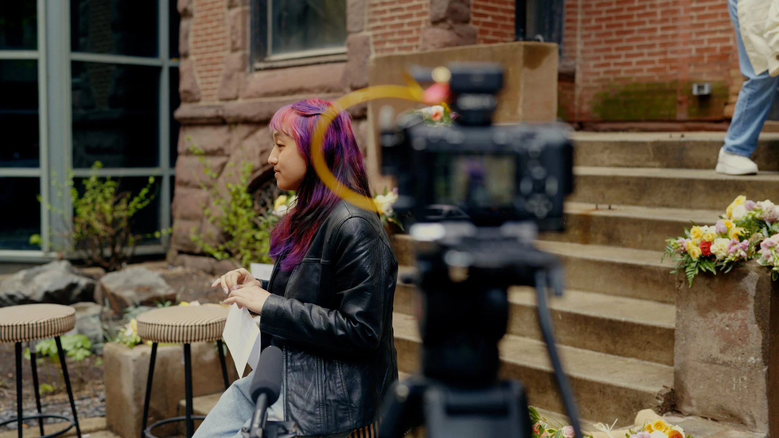 A woman with colorful purple and pink hair wearing a black leather jacket sitting outside near brick steps with flowers, while filming with a camera.