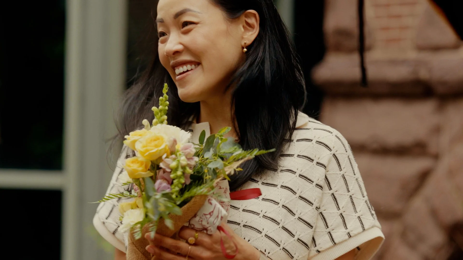 A woman with long black hair, smiling, holding a bouquet of yellow, pink, and purple flowers, wearing a cream-colored sweater with black horizontal stripes, standing in front of a brick wall.