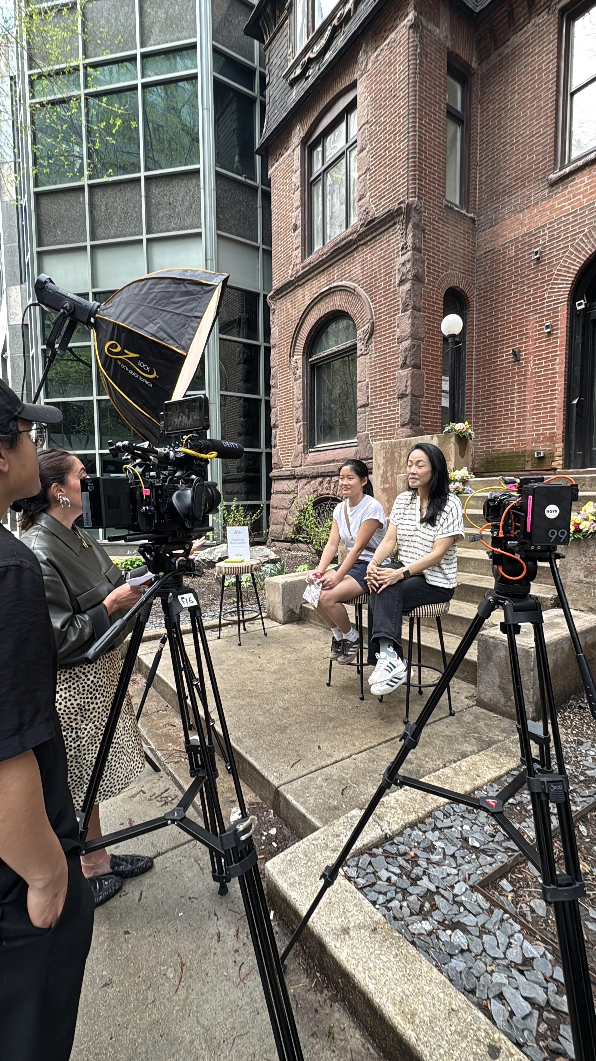 Two women sitting on chairs outdoors, being filmed by a camera crew, in front of a brick building with steps and flowers.