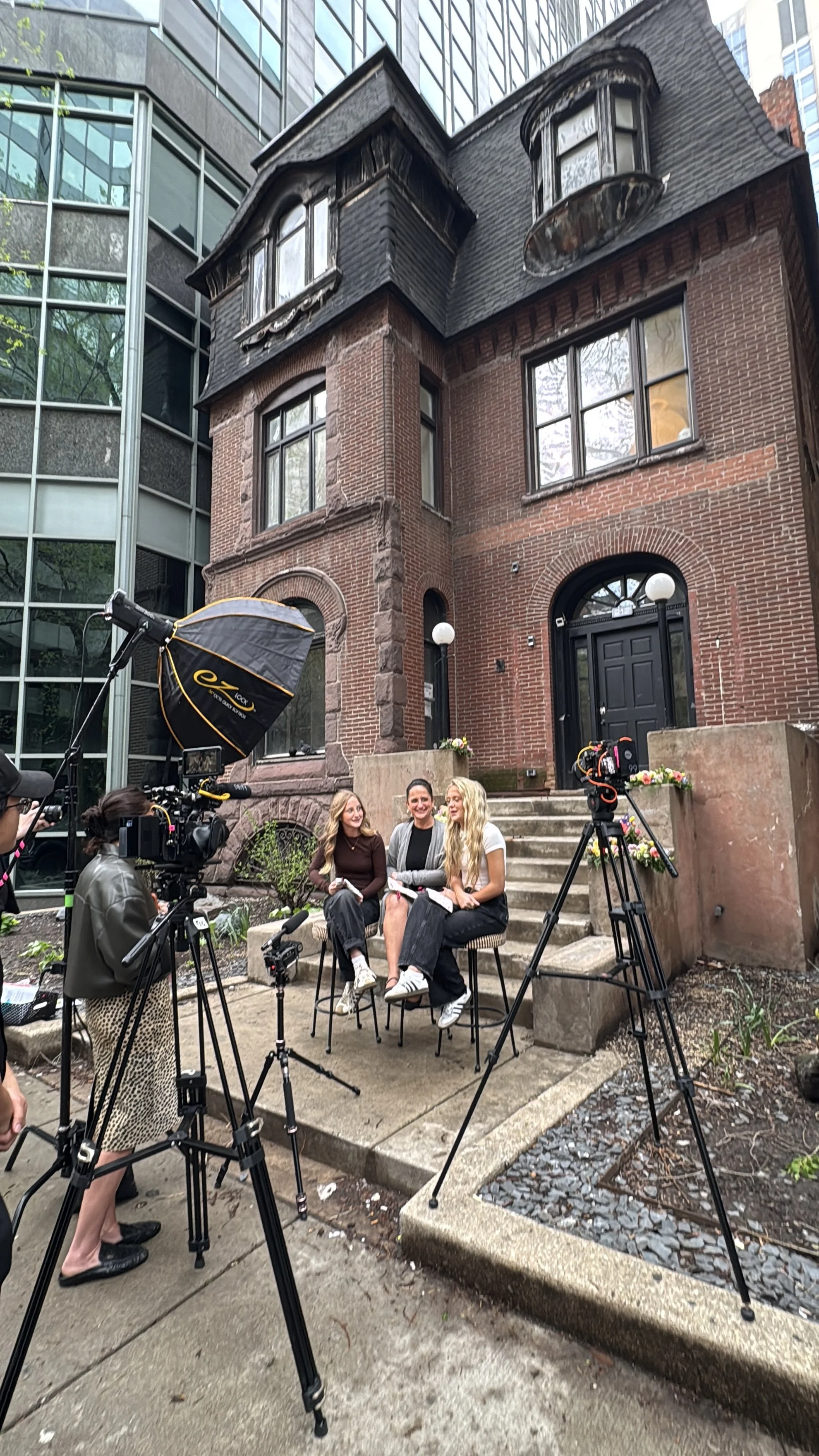 Three women are sitting on chairs outside a brick house, being filmed with multiple cameras and lighting equipment, with a modern glass building in the background.