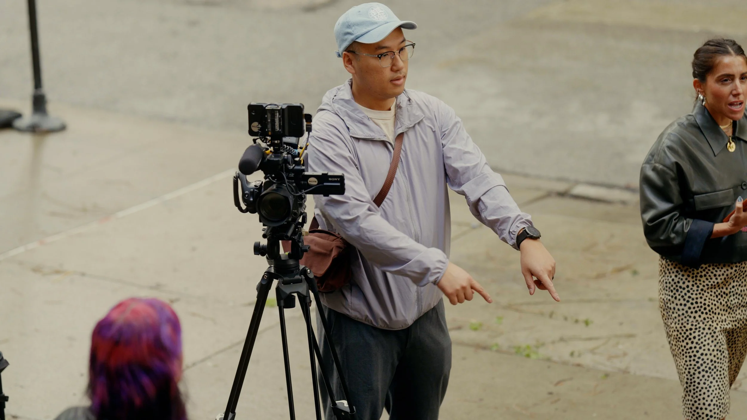 A man in glasses, a light gray jacket, and a baseball cap stands next to a professional camera on a tripod, pointing downwards, as he engages with a woman speaking in front of him during an outdoor event.