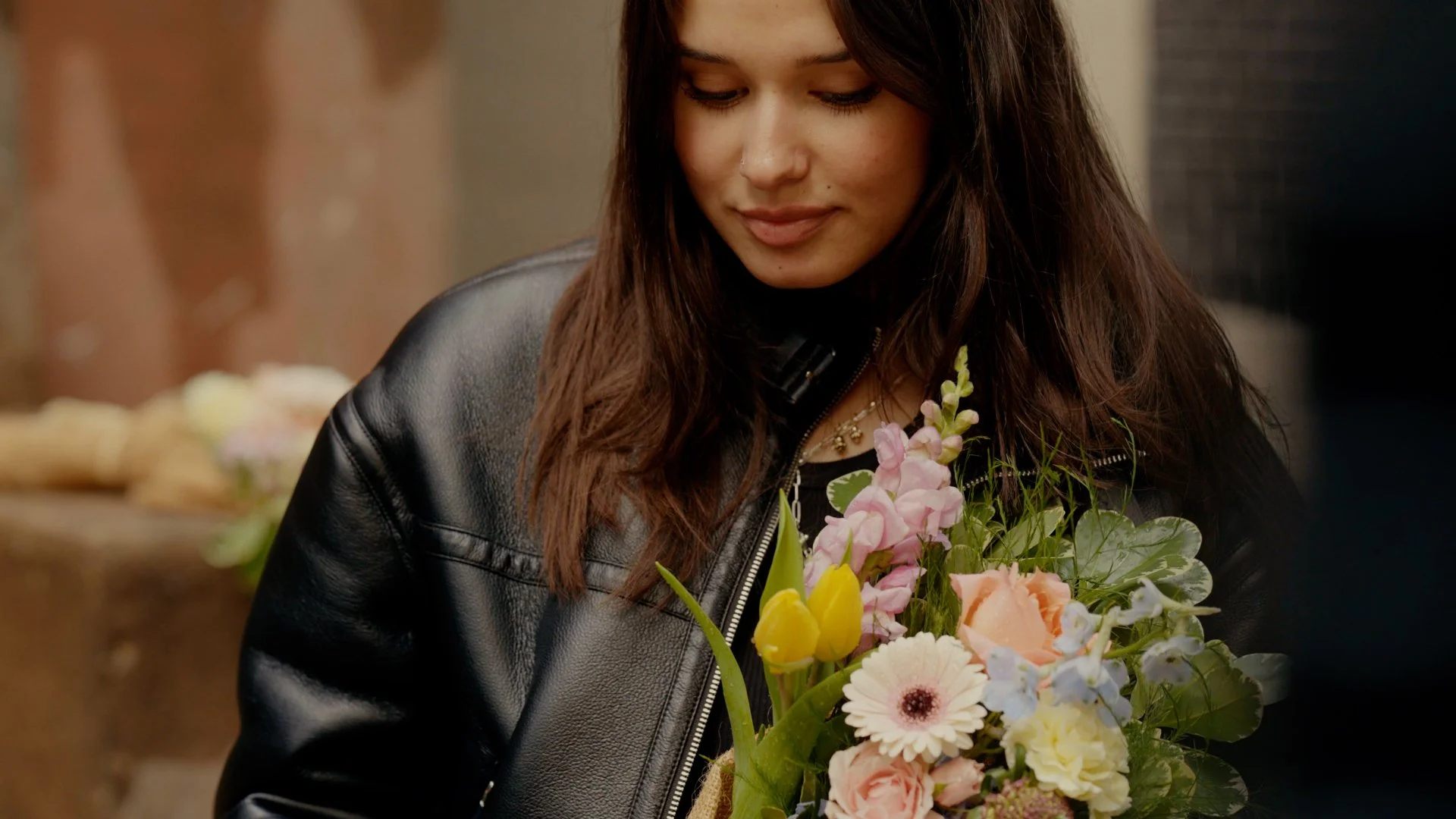 Young woman with brown hair looking down at a bouquet of colorful flowers, wearing a black leather jacket.