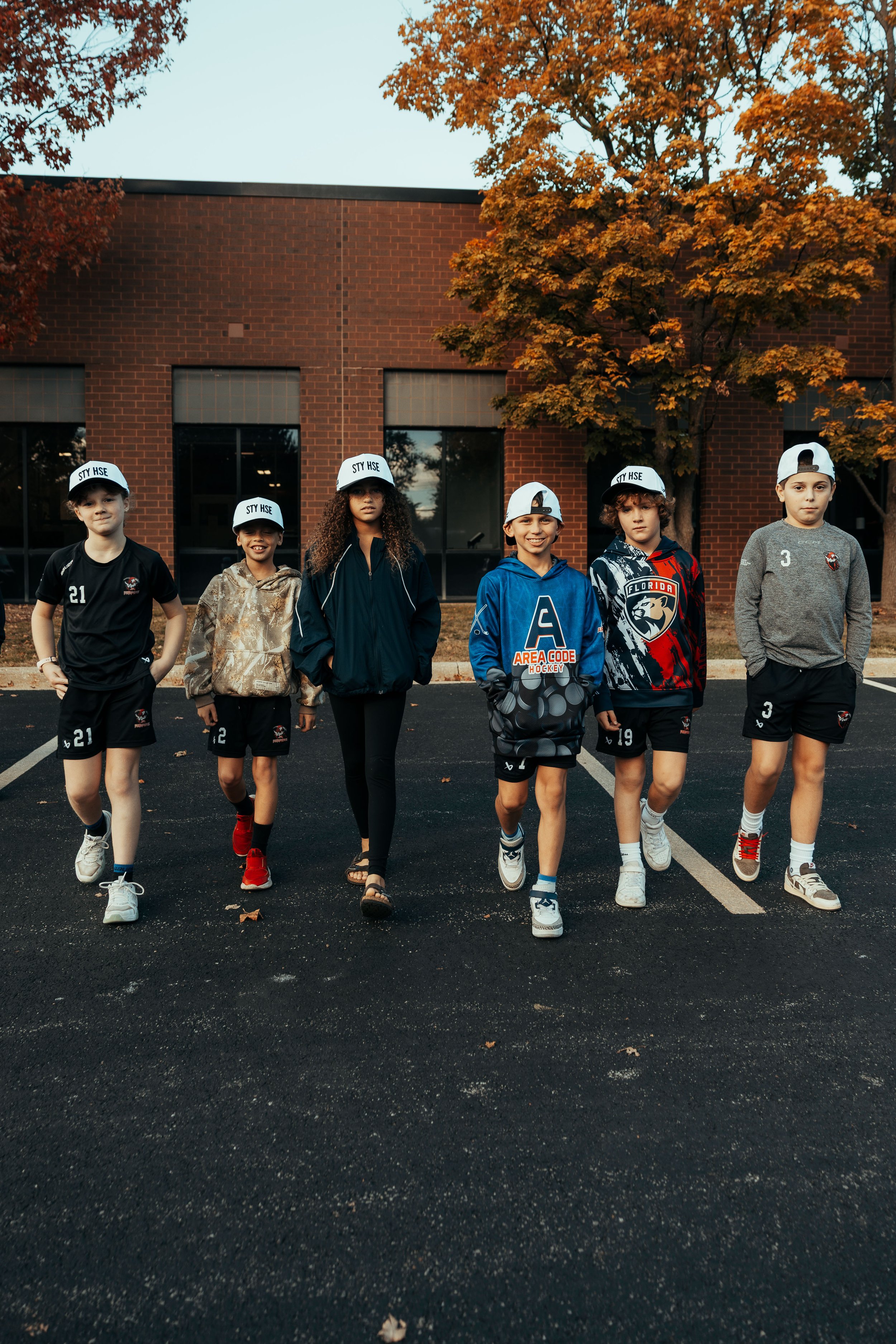 A group of six young kids walking on a parking lot in front of a brick building with trees in the background. The kids are wearing sports apparel and baseball caps.