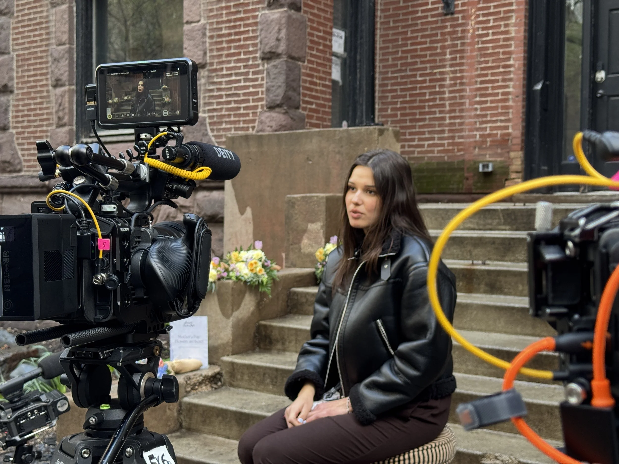 A woman sits on steps outside a brick building during filming, with a professional camera set up to film her. Flowers are arranged nearby.