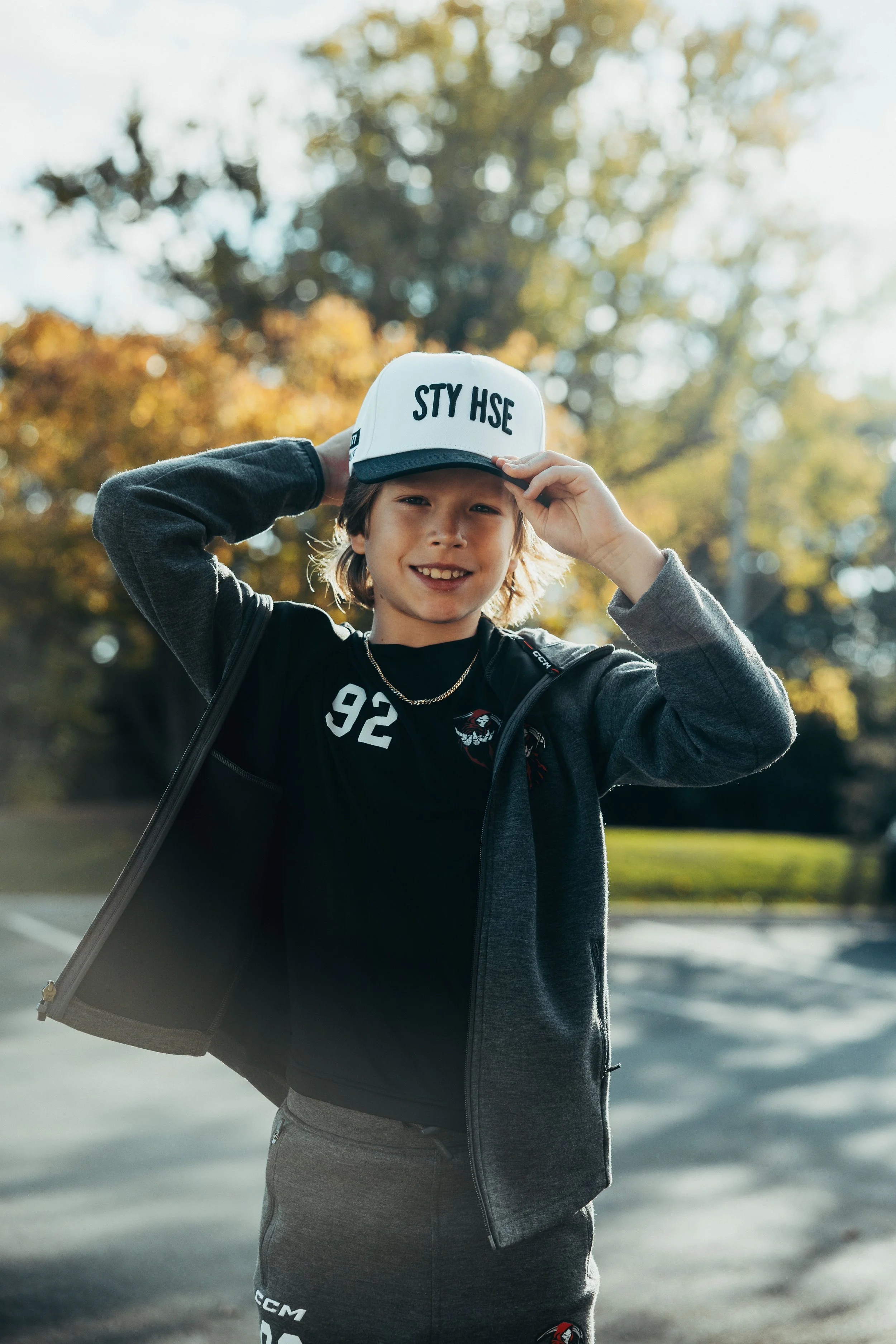A young boy outdoors wearing a white cap with the phrase 'STY HSE' and a black Montreal Canadiens hockey jersey, adjusting his cap with a smile, with autumn trees in the background.
