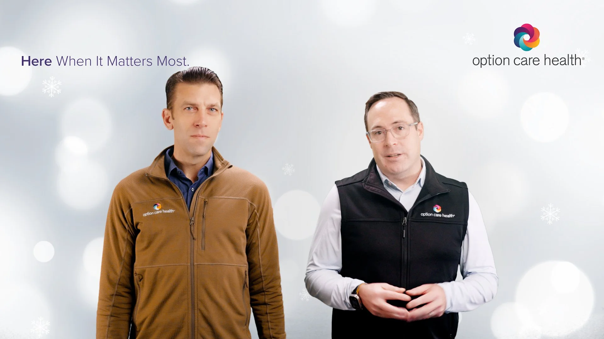 Two men wearing jackets with the Option Care Health logo standing against a white background with snowflakes. One wears a brown jacket, the other a black vest.
