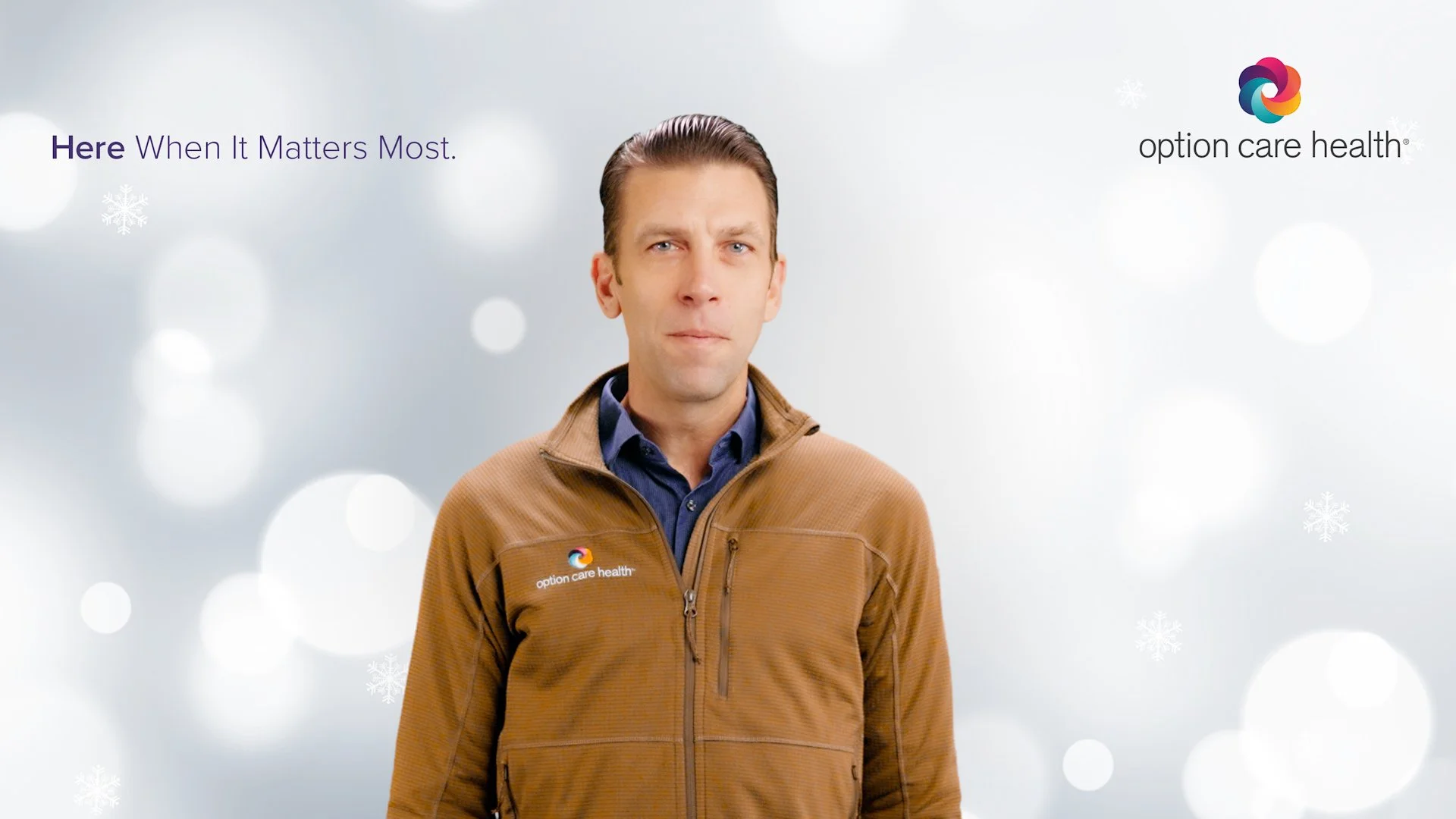 A man wearing a brown jacket with a logo that reads 'option care health' on the chest stands against a festive, snowy background with snowflakes and bokeh lights, looking at the camera.