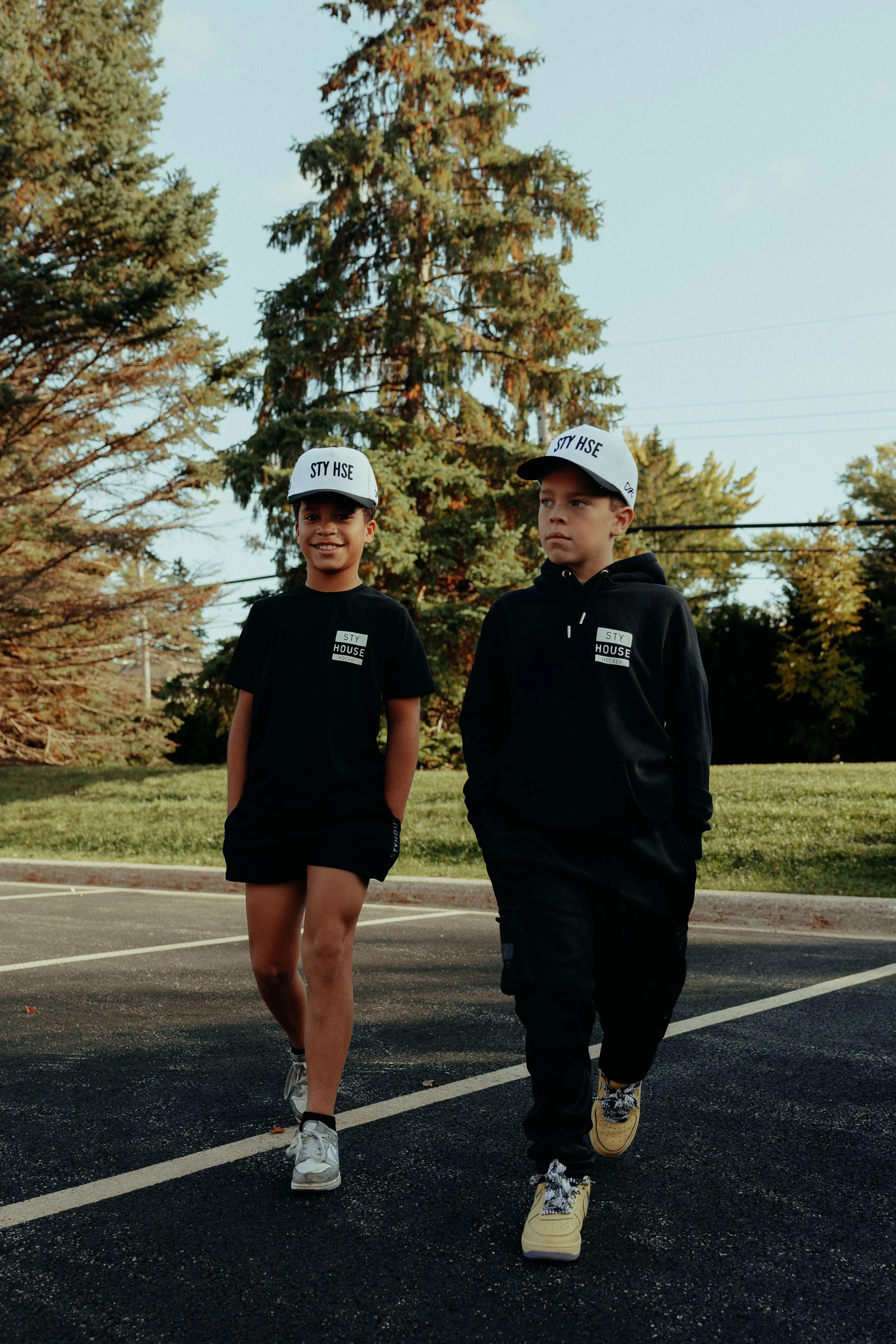 Two young teenagers walking in a parking lot with trees and a clear blue sky in the background. Both are wearing black hoodies and white caps with black text, and casual footwear. The girl is smiling and the boy has a serious expression.