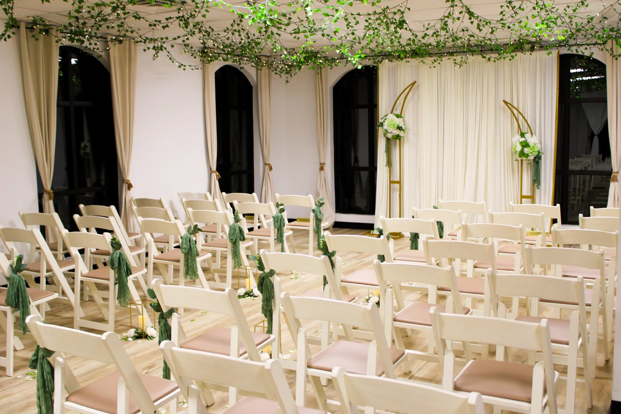 Wedding ceremony setup with white chairs, green decorations, and floral arrangements against a white curtain backdrop.