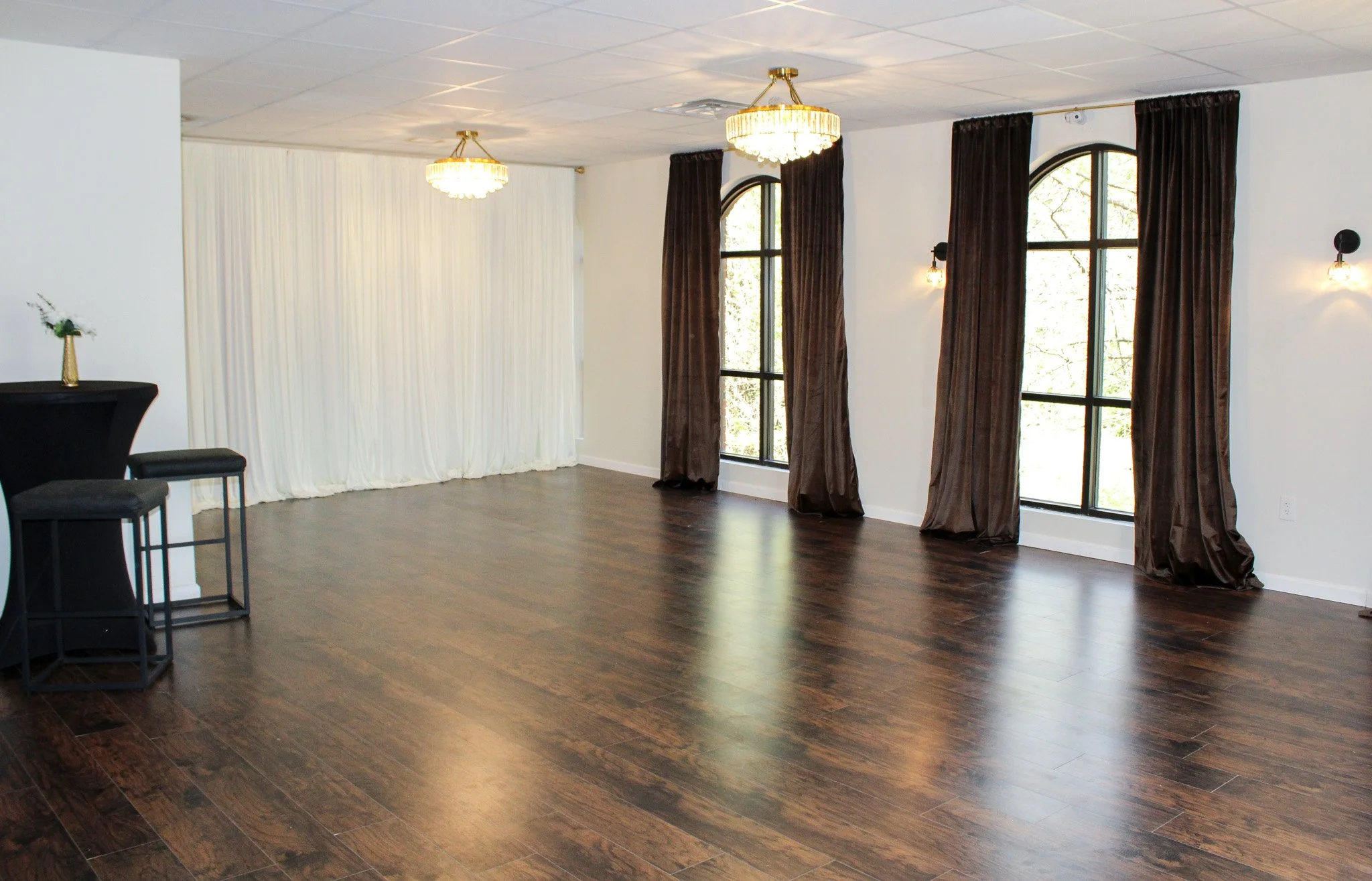 Empty banquet hall with dark wood floors, white walls, large arched windows with dark curtains, and chandeliers hanging from the ceiling.