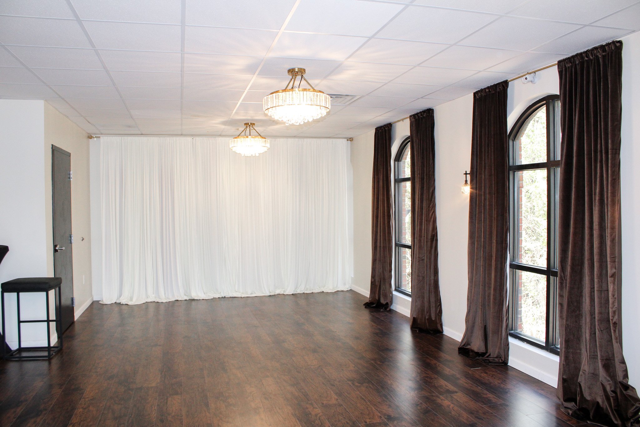 Empty event or wedding venue with dark wood flooring, white walls, large windows with dark curtains, white draped backdrop, and chandeliers.