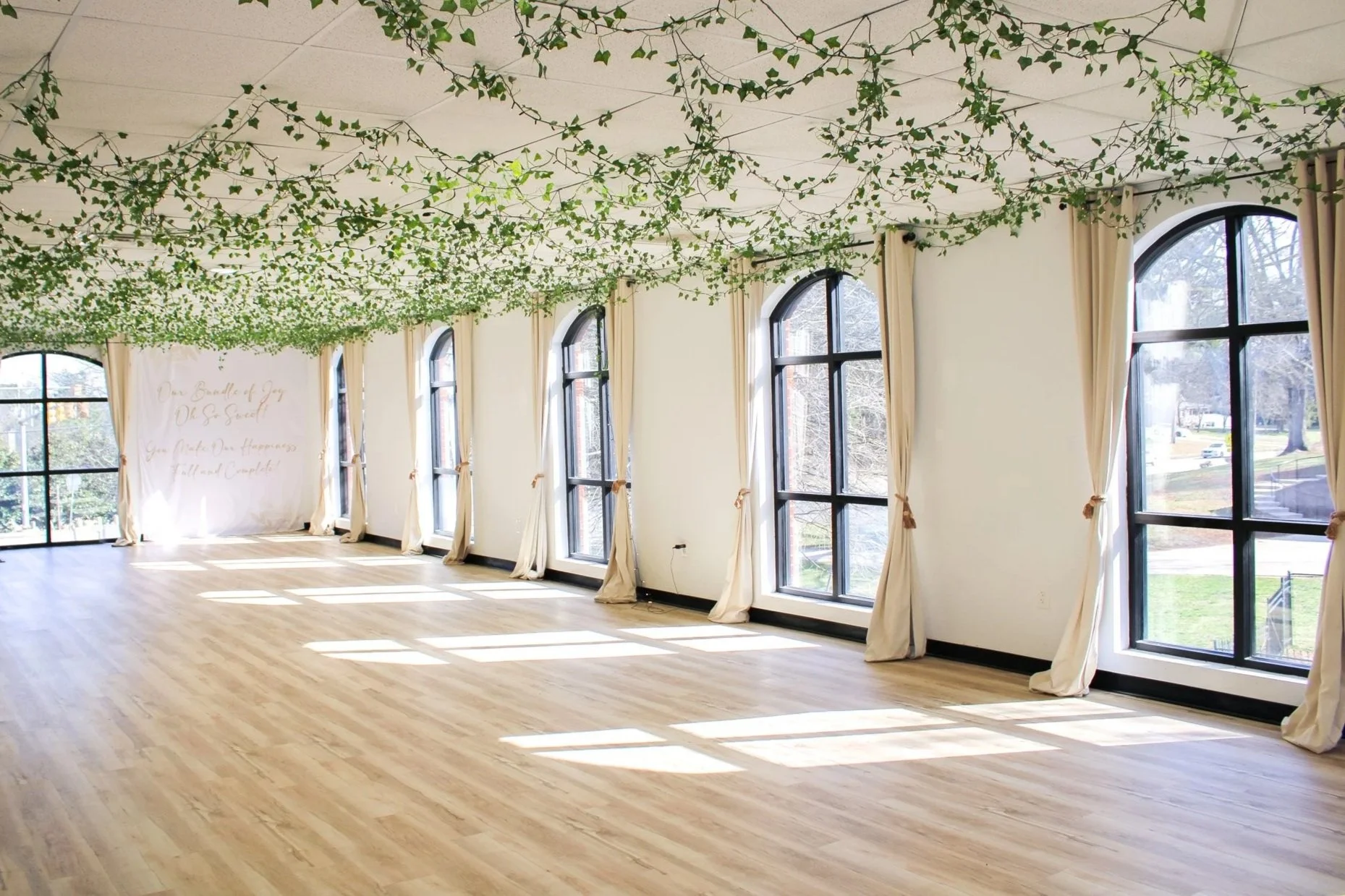 A spacious room with large arched windows allowing natural light, decorated with flowing cream curtains tied back, featuring green leaf garlands hanging from the ceiling, and a light wood floor. Outside, trees and a grassy area are visible.
