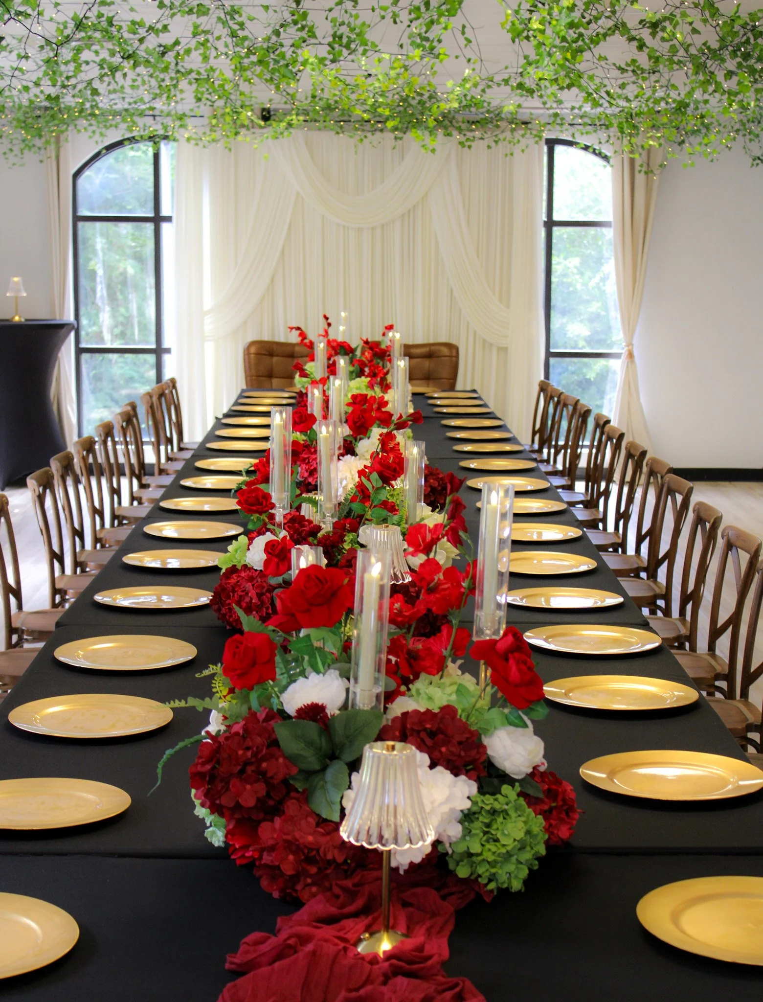 Elegant banquet table setup with a black tablecloth, yellow plates, and a floral centerpiece of red, white, and green flowers. There are candles and decorative lamps on the table. The room has large windows with white curtains and greenery outside, and a ceiling decorated with green foliage and string lights.