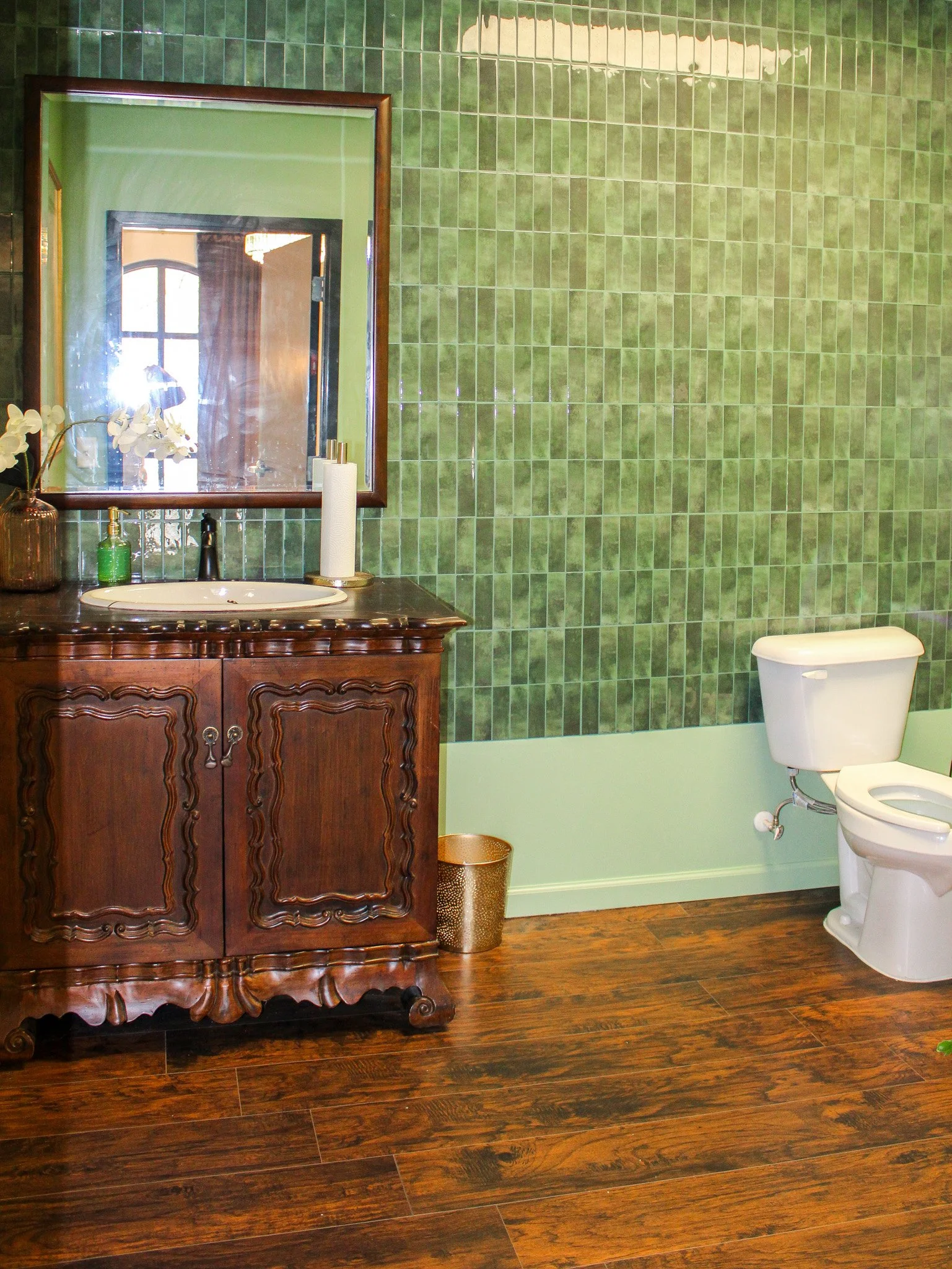 Bathroom with green tiled wall, wooden vanity with a mirror, white toilet, and dark wood flooring.