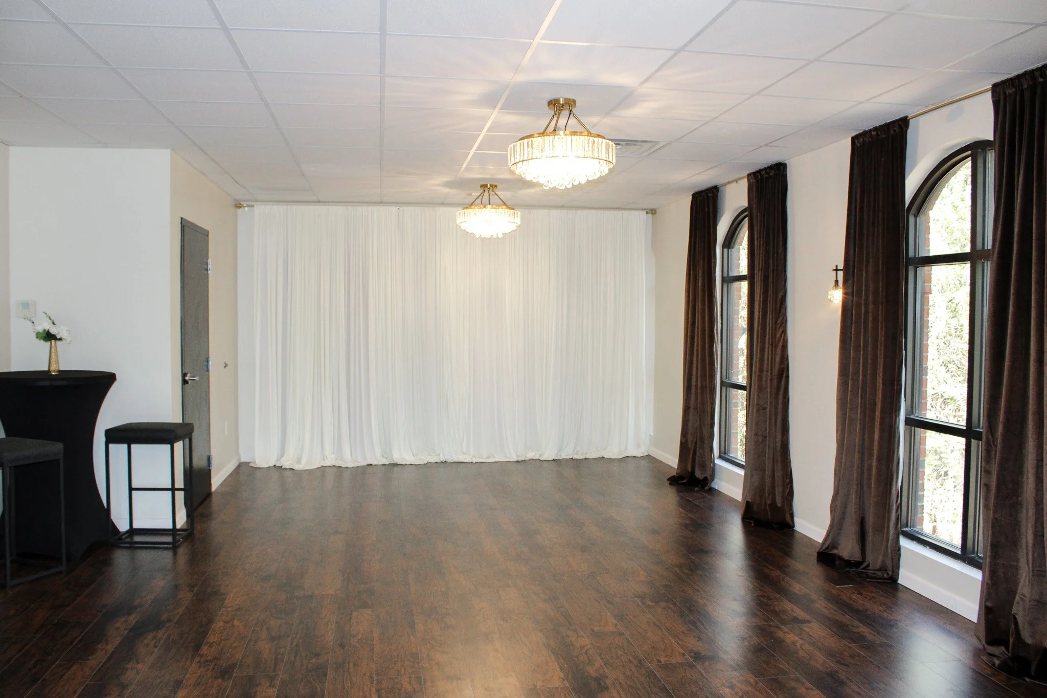 Empty event space with dark wood floor, white walls, large window with brown curtains, and a white draped backdrop at the far end. Two chandeliers hang from the ceiling, and there is a small black table with a flower arrangement and two stools on the