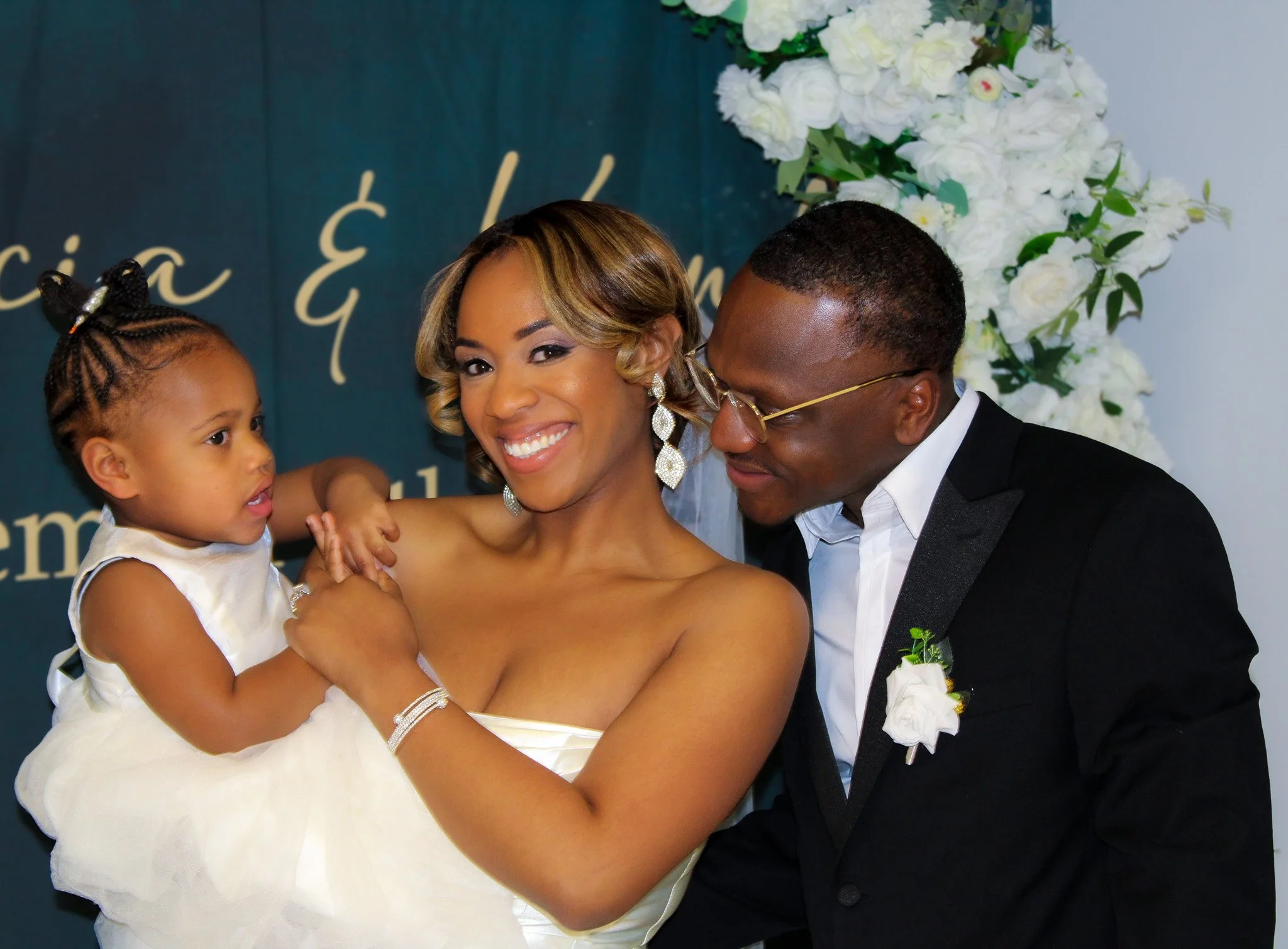 A smiling woman holding a young girl, with a man looking at them, in front of a floral backdrop and a dark green background with decorative writing. The woman is wearing earrings and a strapless dress, the man is wearing glasses, a suit, and a boutonnière, and the girl is wearing a cream dress with braided hair.