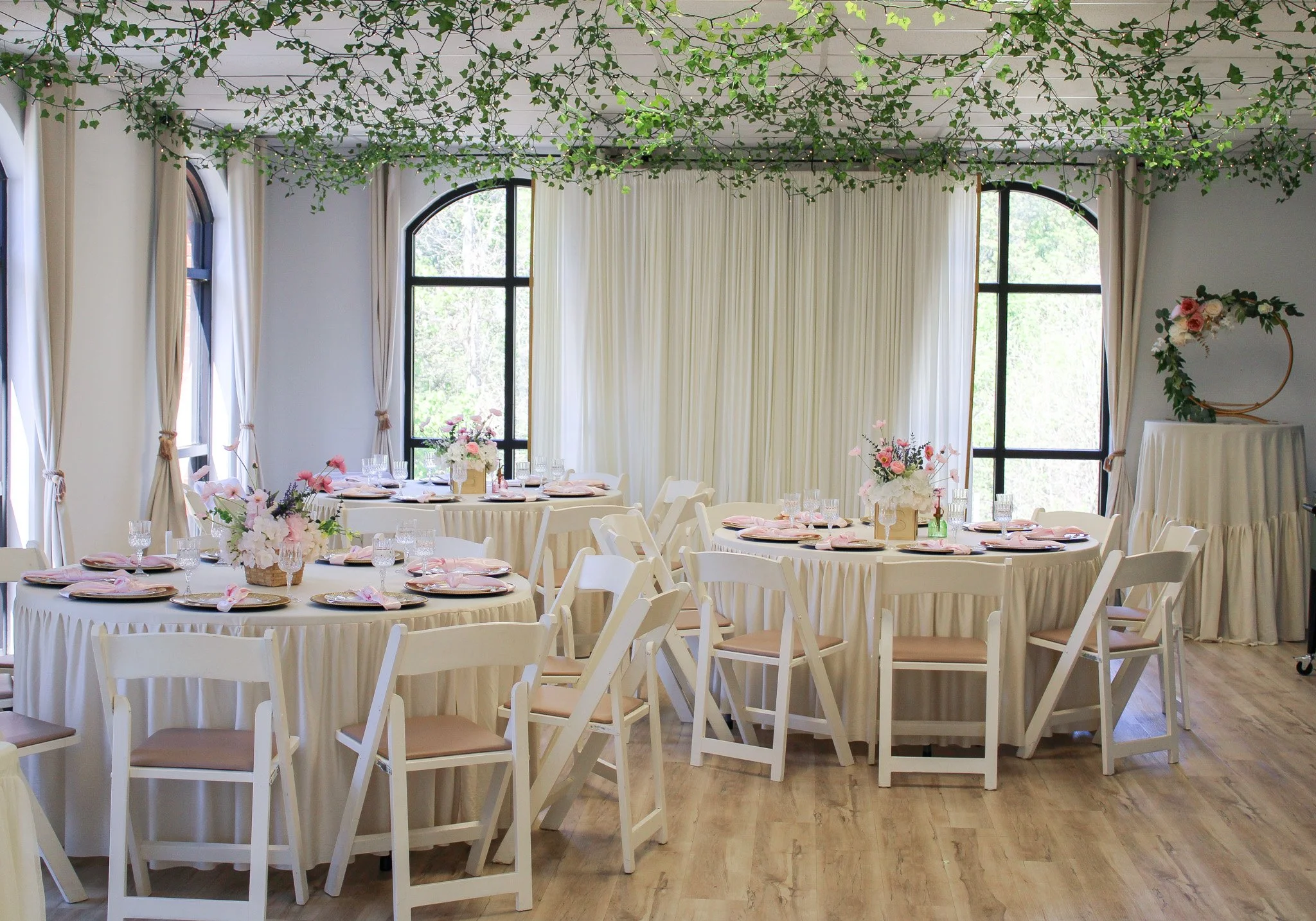 Elegant dining setup with round tables covered in cream tablecloths, decorated with pink and white floral centerpieces, set with pink napkins, plates, and glasses. Surrounded by white chairs with beige cushions. The room features large arched windows