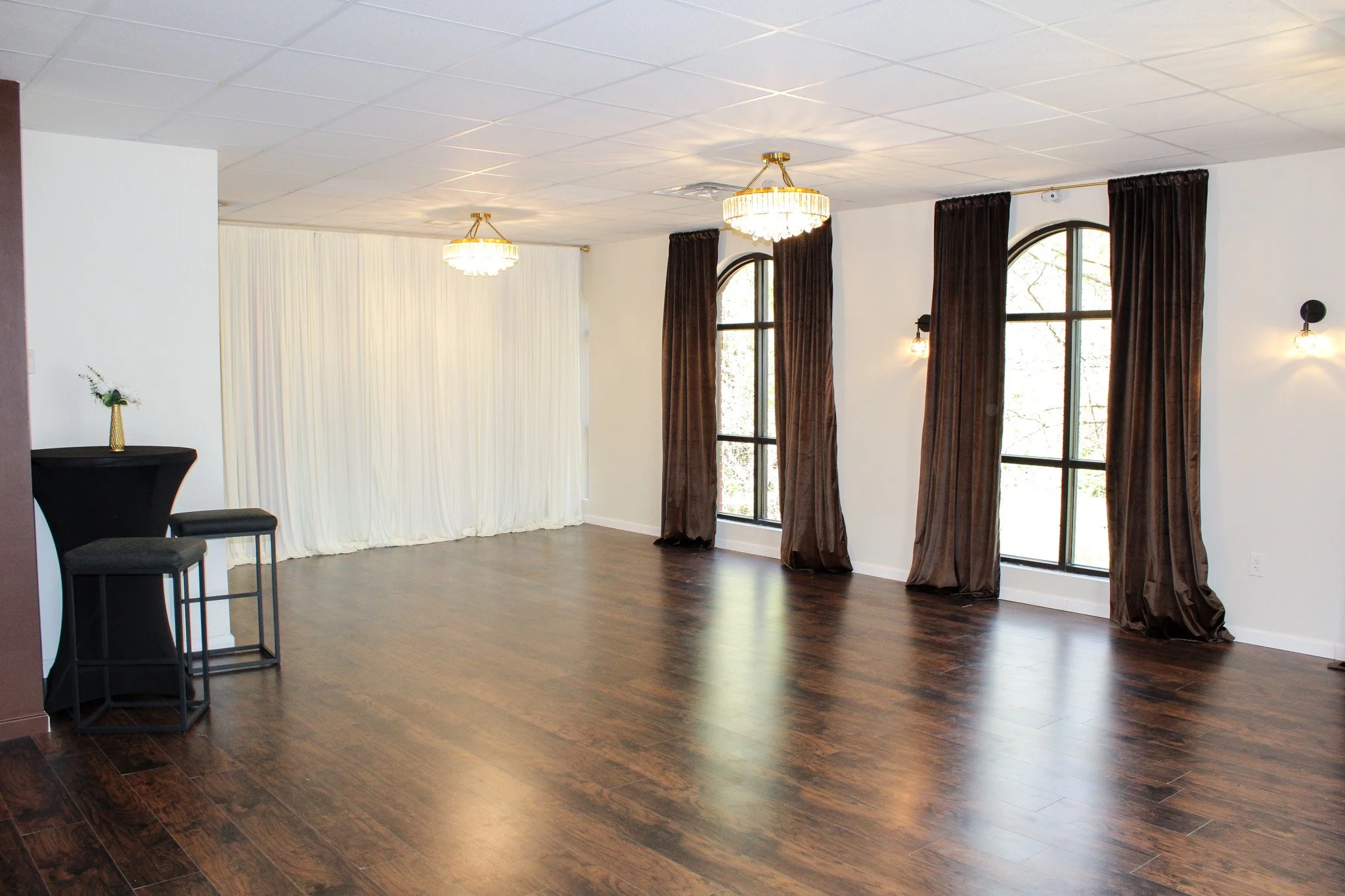 Empty event space with wooden floors, large arched windows with dark curtains, white walls, and chandeliers hanging from the ceiling.