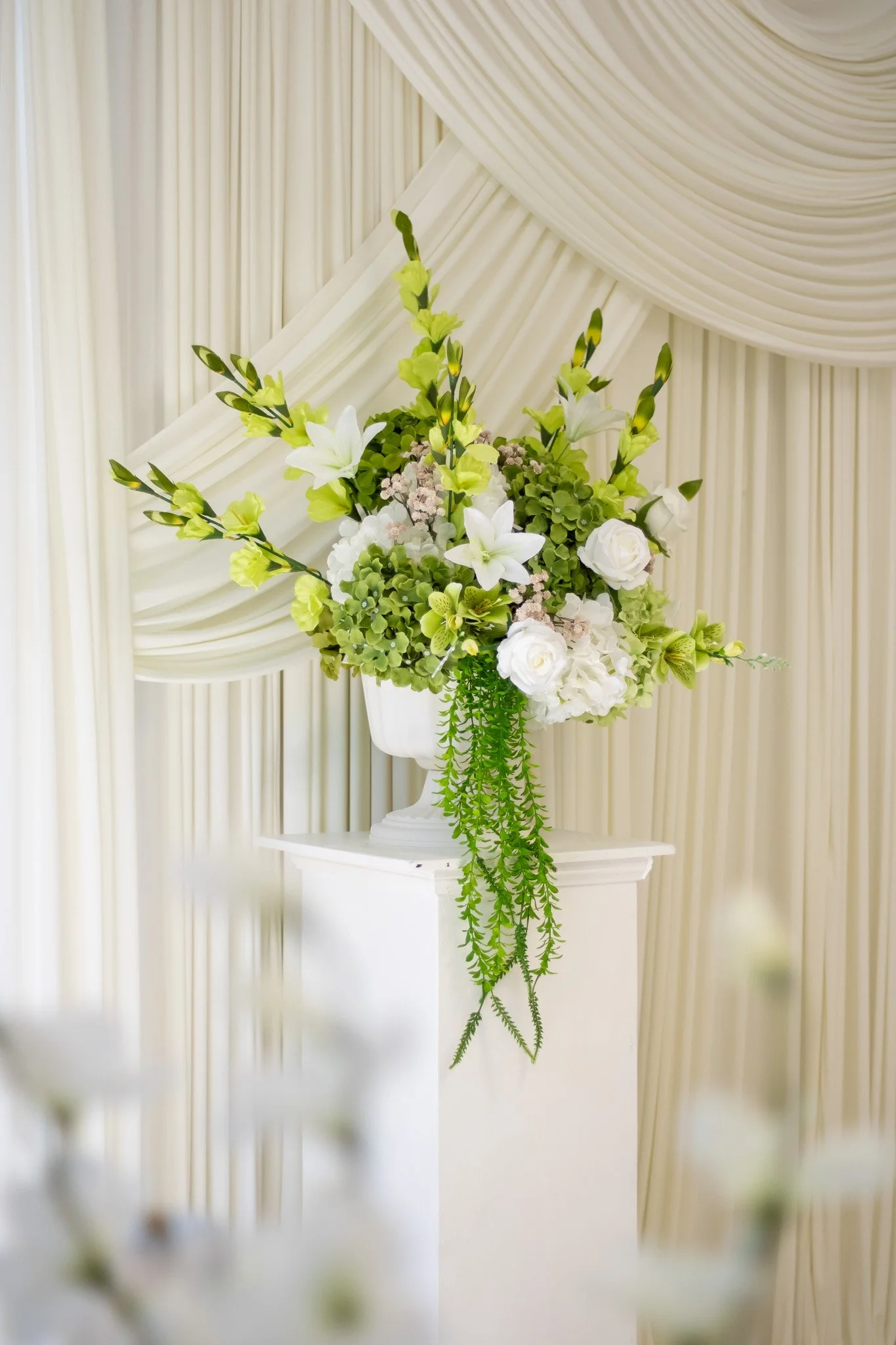 A floral arrangement with white lilies, roses, and green foliage in a white vase, placed on a white pedestal in front of cream-colored draped curtains.