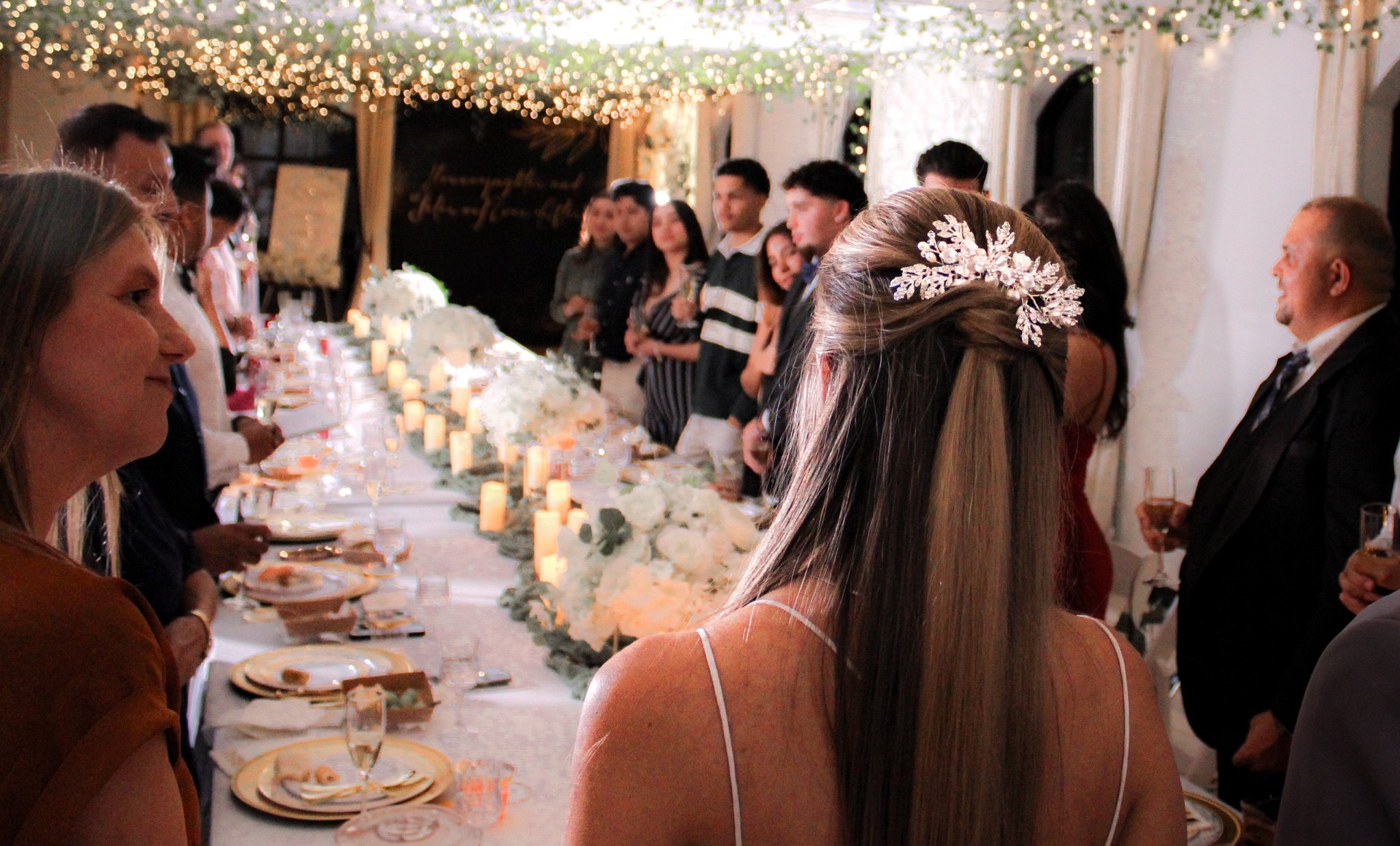 A wedding reception with a long decorated table, floral centerpieces, candles, and guests standing and clinking glasses, with a bride wearing a floral hairpiece in the foreground.
