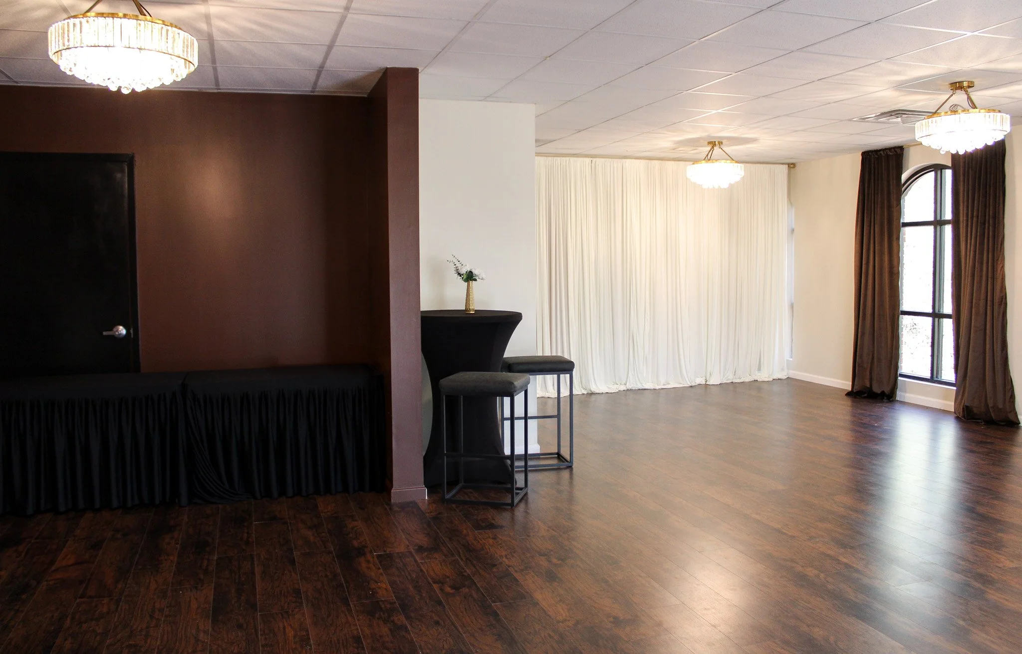 Empty event room with hardwood floors, white ceiling, dark curtains, and large windows, illuminated by hanging chandeliers, with a small high-top table and two stools near a decorative curtain.