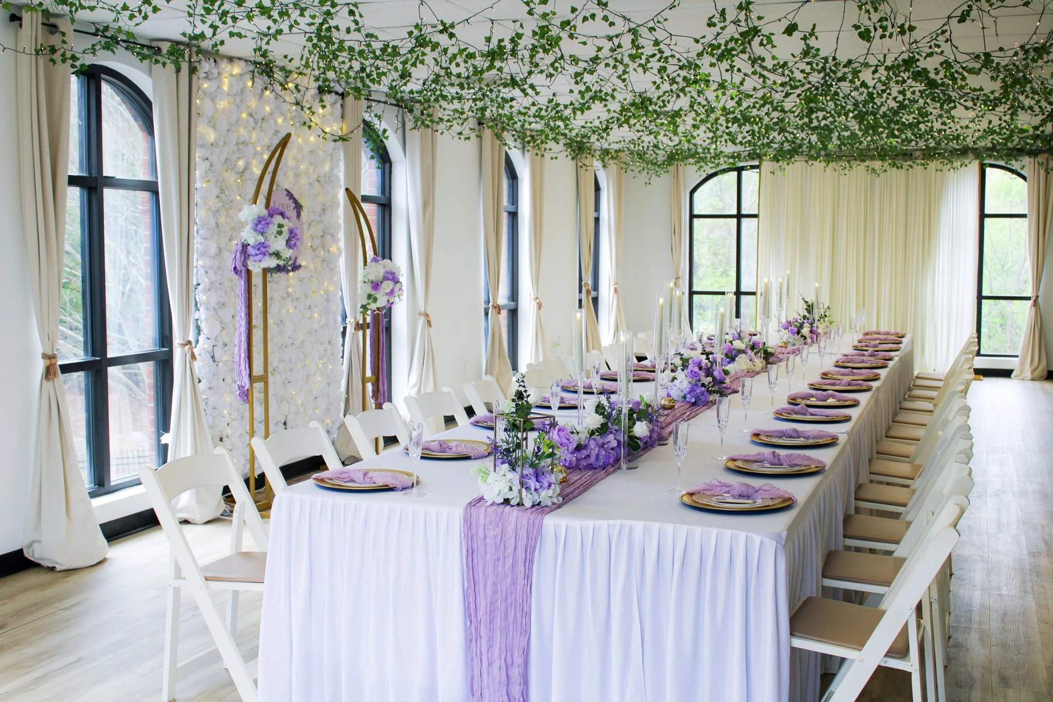 Elegant banquet table decorated with purple and white flowers, tall candles, and lavender fabric, set in a bright room with large windows, cream curtains, and green ceiling decorations.