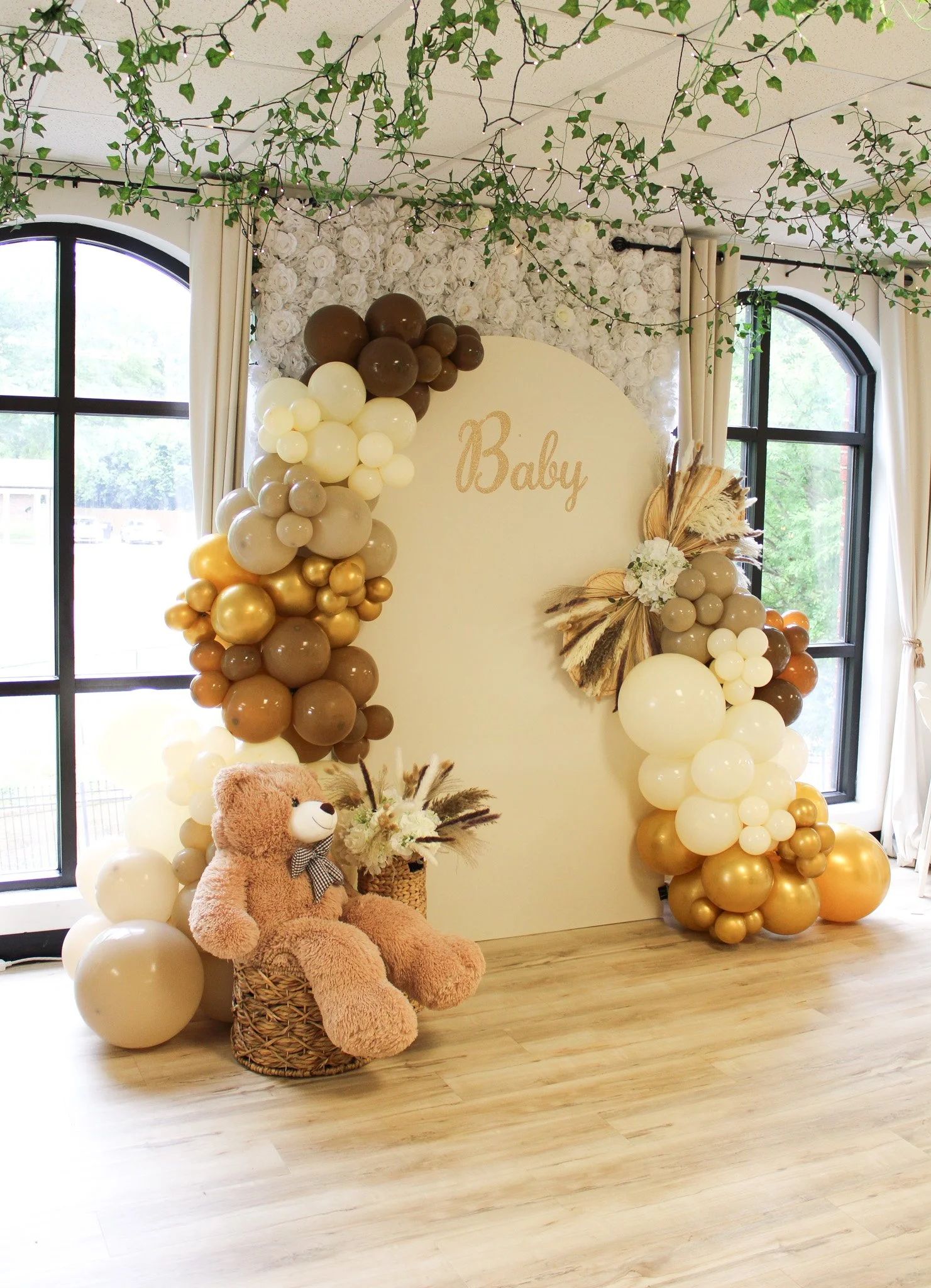 Decorative setup for a baby shower with pink, white, and gold balloons, floral arch, sign that says 'Oh Baby,' cake with 'Oh Baby' topper, and a table with a sign that says 'Sweet'.