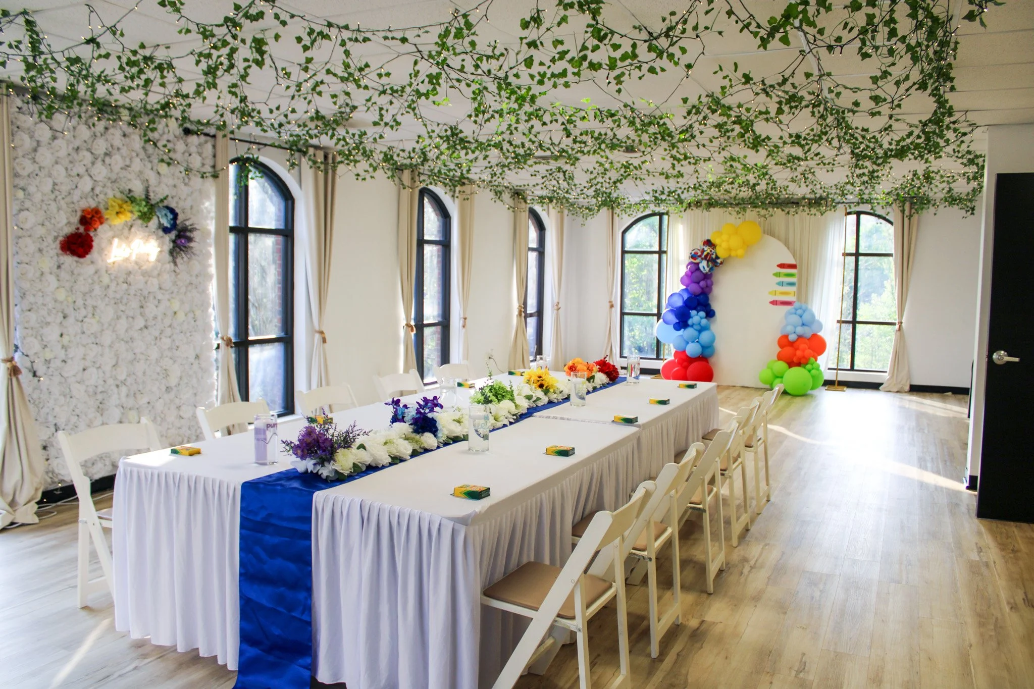 Decorated indoor event space with a long white table with floral centerpieces, surrounded by white chairs, and colorful balloon arch at the far end near tall windows. Green garland with string lights hangs from the ceiling.