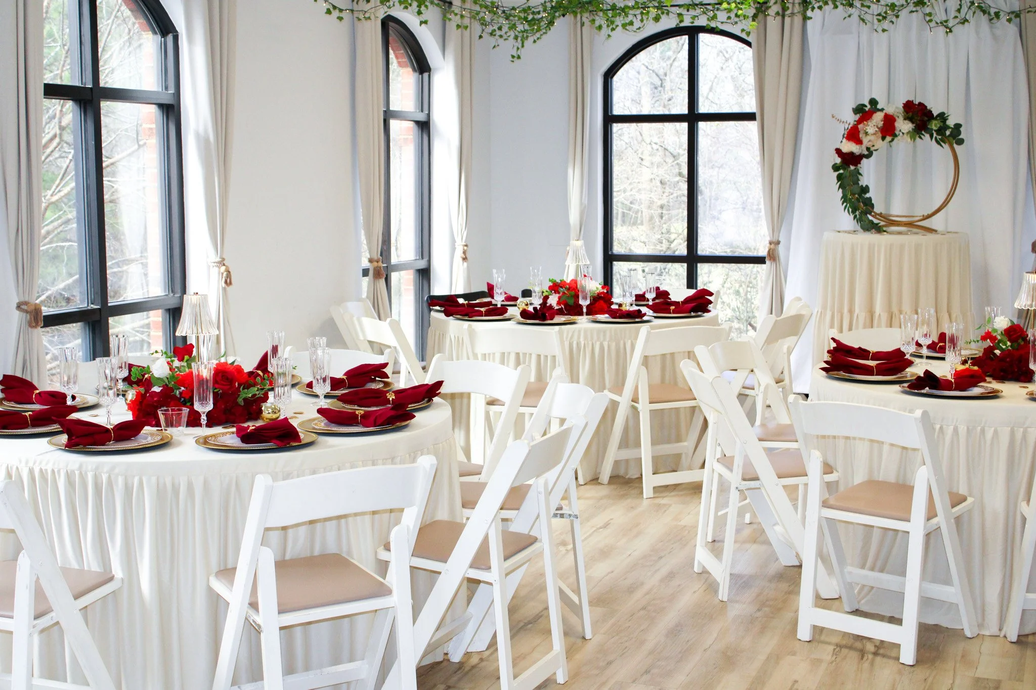 Wedding decorated reception room with white tables, red floral centerpieces, red napkins, and white chairs, large windows with white curtains, and a floral hoop decoration on a stand.