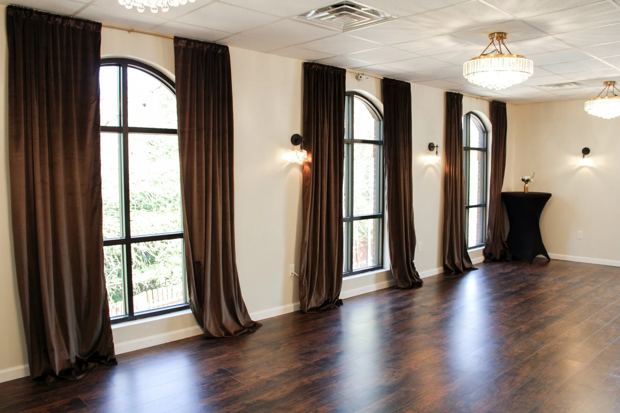 An empty room with four large arched windows with dark curtains, dark wood flooring, wall-mounted lights, and a black table with a flower vase in the corner.