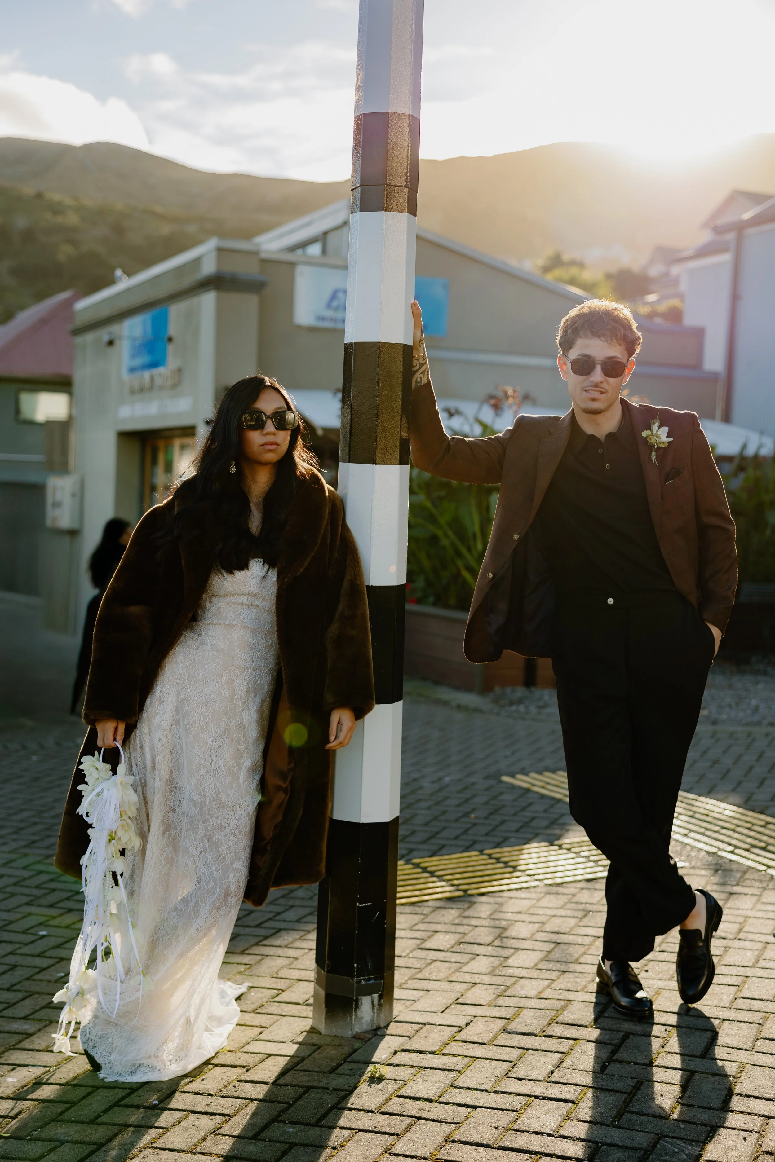 A young woman and a young man pose outdoors in front of a striped pole during sunset. The woman wears a white lace dress, a brown fur coat, and sunglasses, holding a bouquet of white flowers. The man wears a black outfit, sunglasses, and a suit jacke