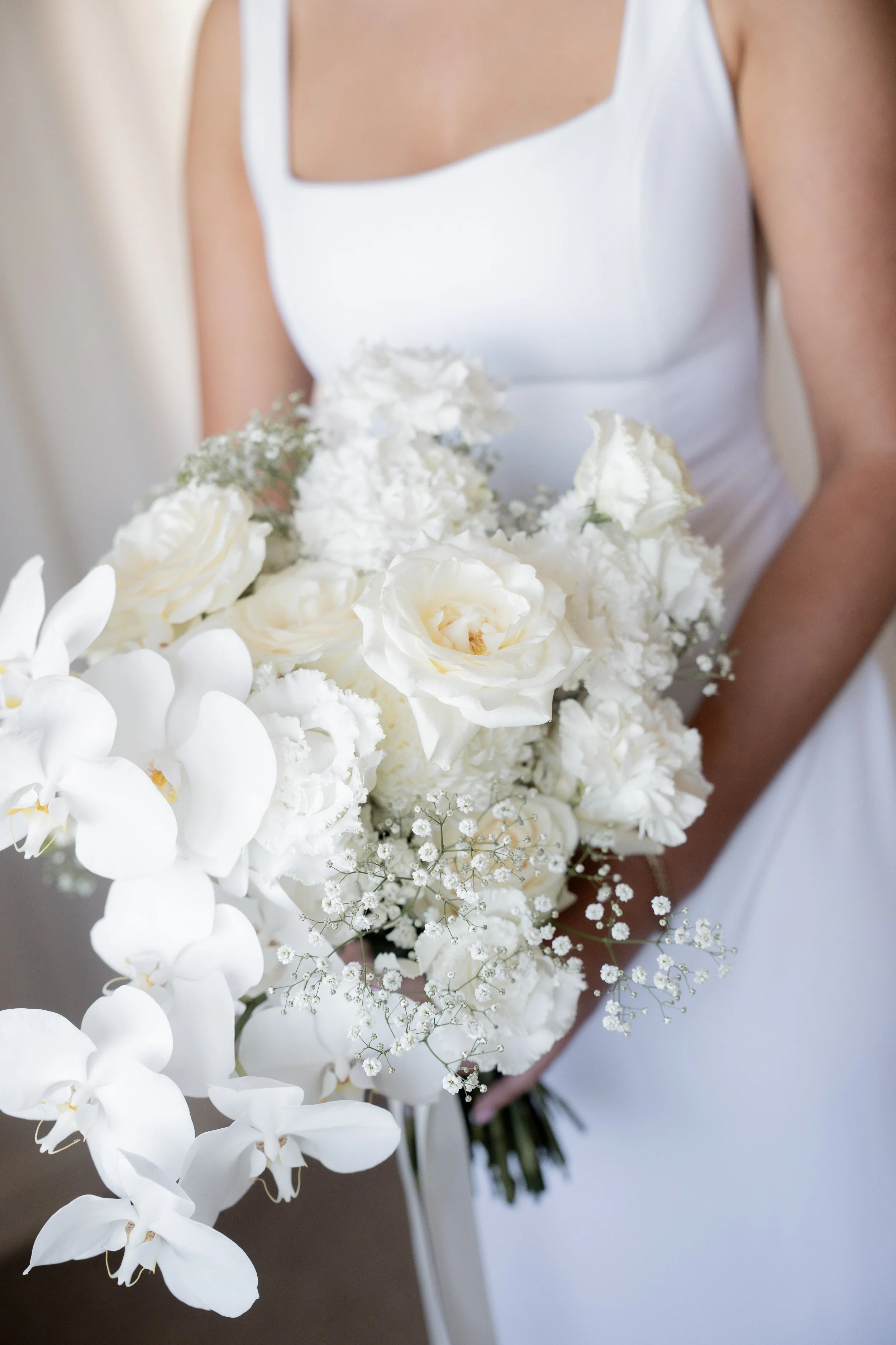 A person wearing a white dress is holding a bouquet of white flowers, including orchids, roses, and baby's breath.