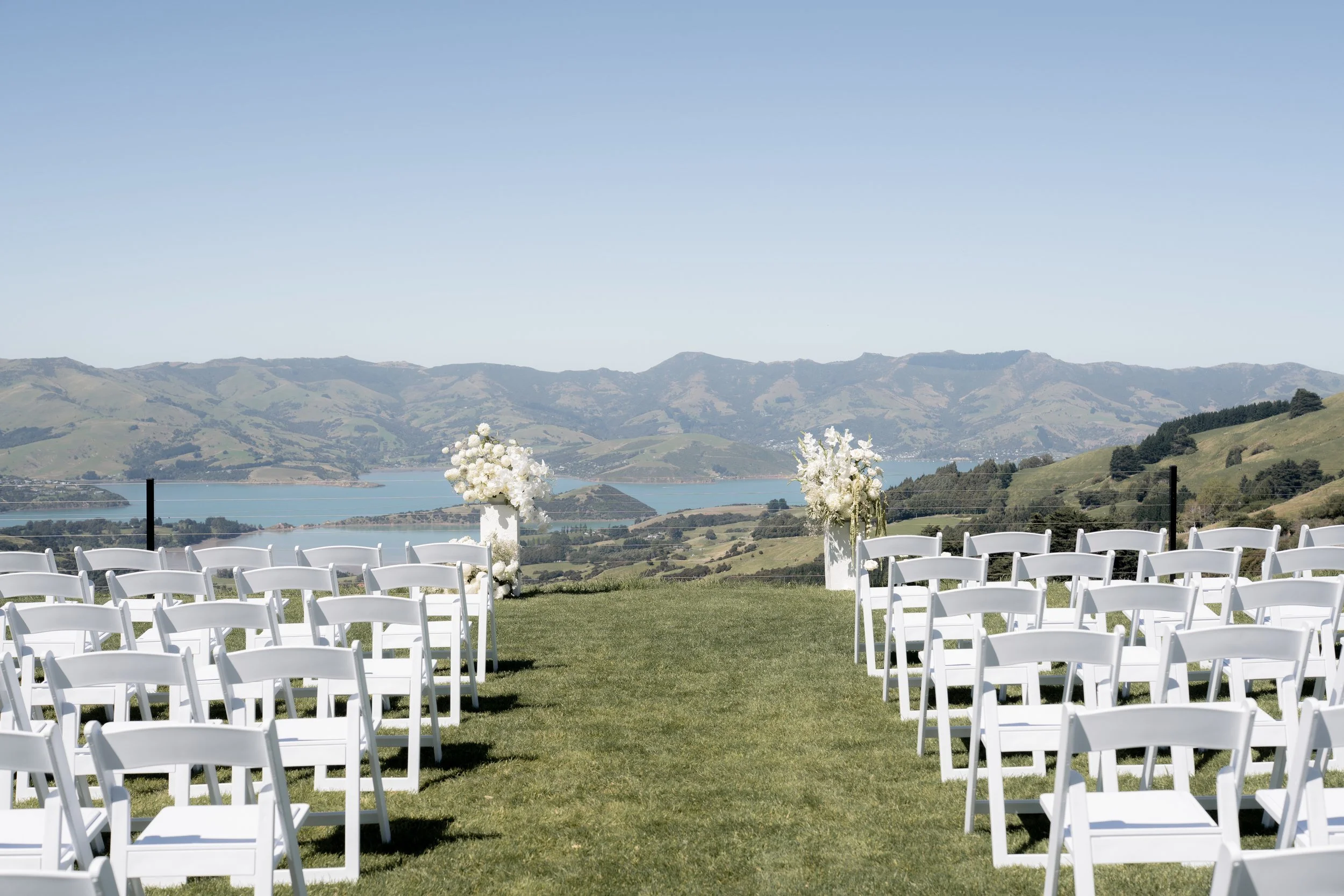 Outdoor wedding setup with white chairs arranged on a grassy field facing floral arrangements, overlooking a body of water and mountains under a clear blue sky.