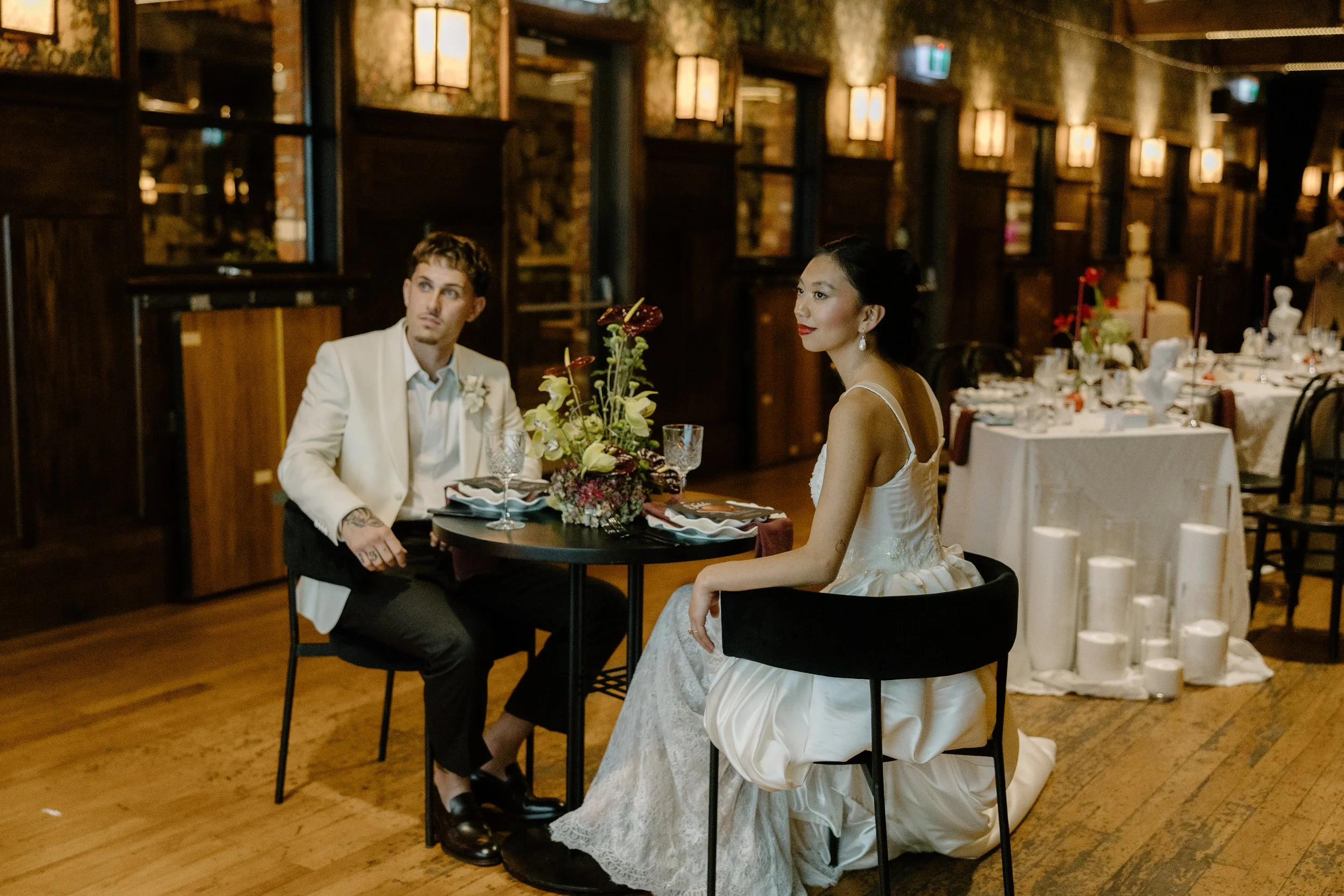 A bride and groom sit at a table in a warmly lit restaurant or banquet hall, with the bride wearing a white wedding dress and the groom in a white suit jacket. The table is decorated with a floral centerpiece and glassware, and the background feature