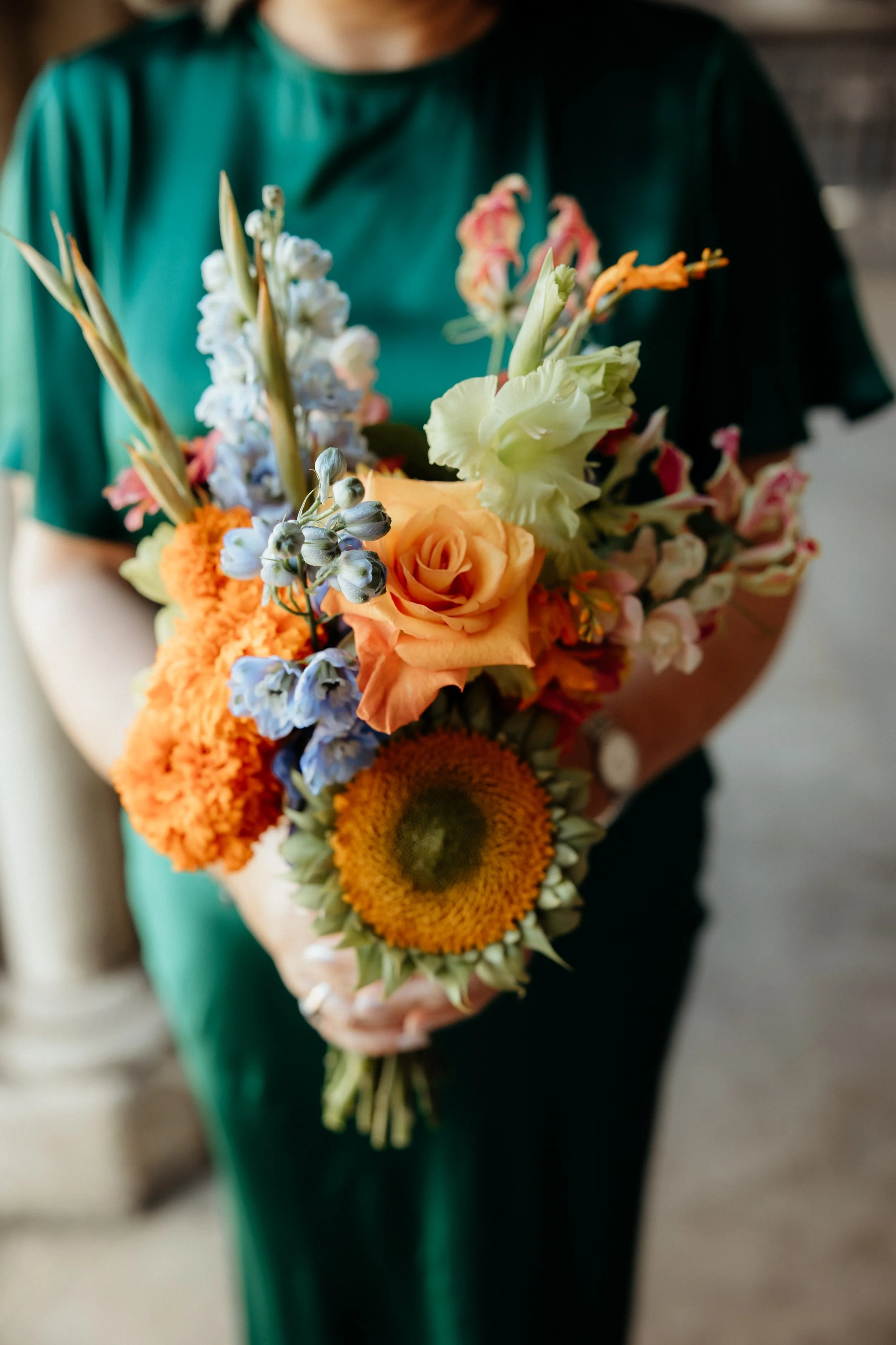 Person wearing a teal dress holding a bouquet of mixed flowers including orange roses, white lilies, blue delphiniums, and sunflowers.