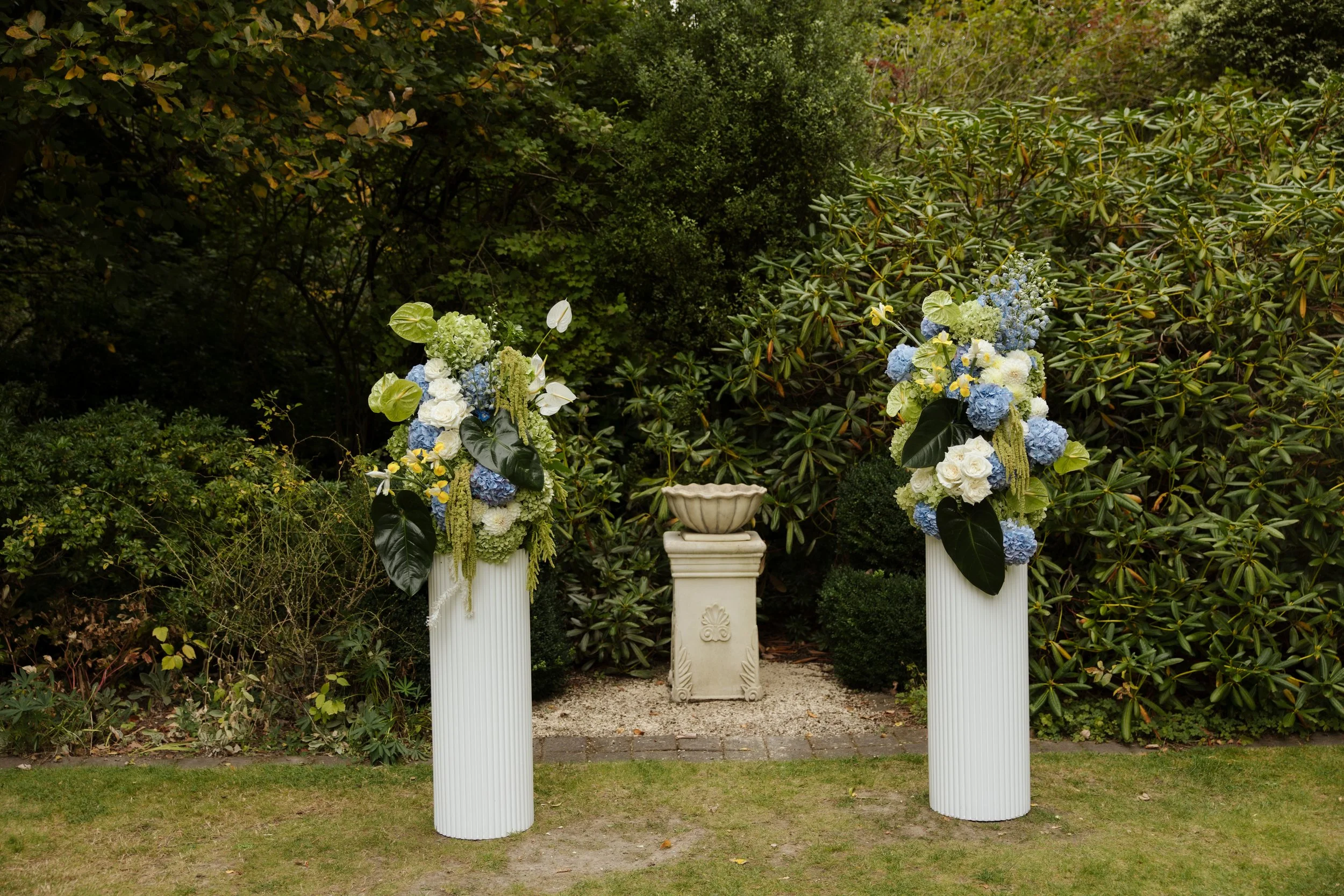 Outdoor garden scene with two white columns decorated with flowers, green leaves, and yellow accents, and a third smaller pedestal with a decorative bowl, surrounded by lush green bushes and trees.