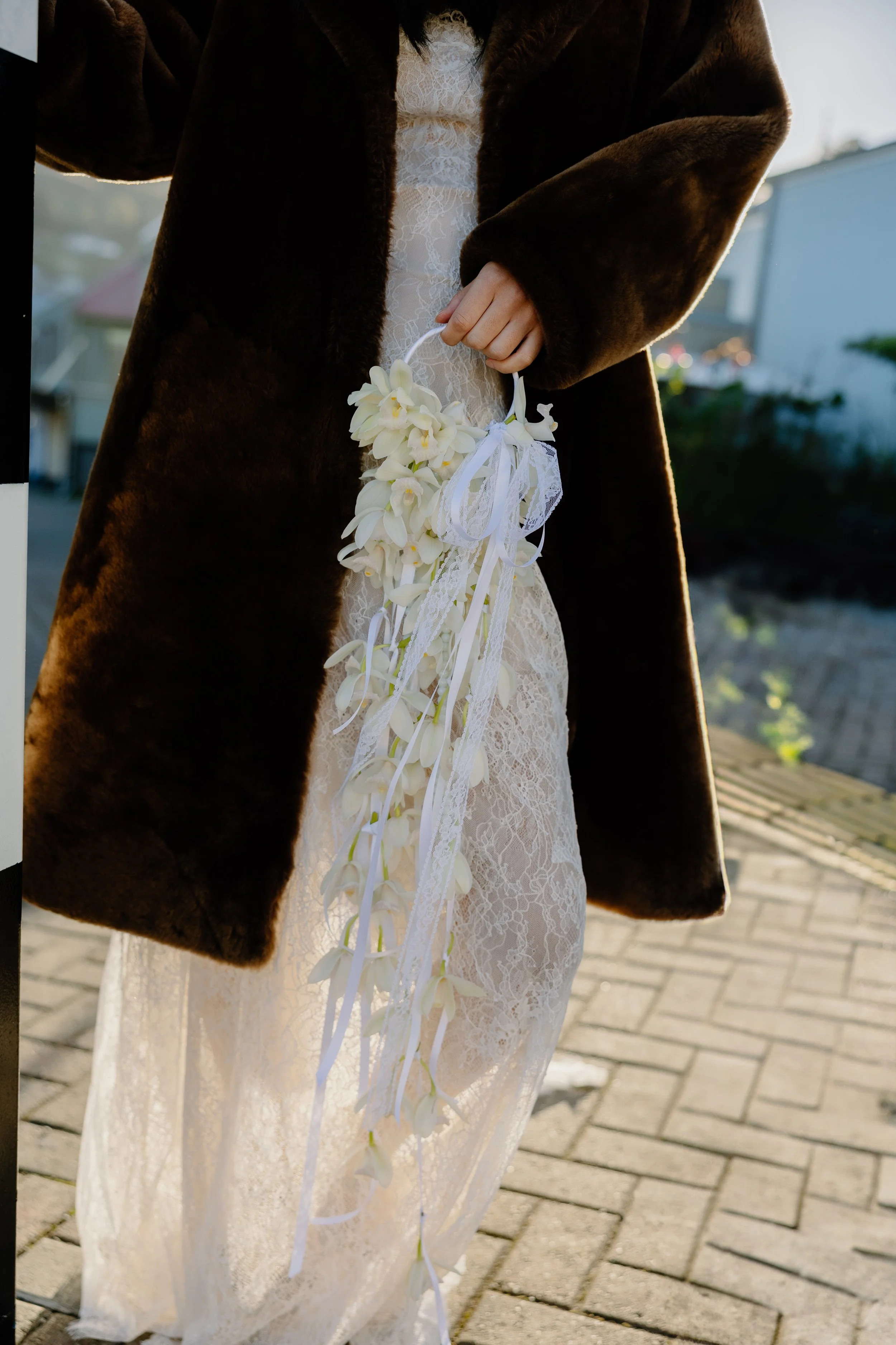 A woman in a white lace dress holding a hanging floral arrangement with white flowers and ribbons, wearing a dark fur coat, outdoors in natural lighting.