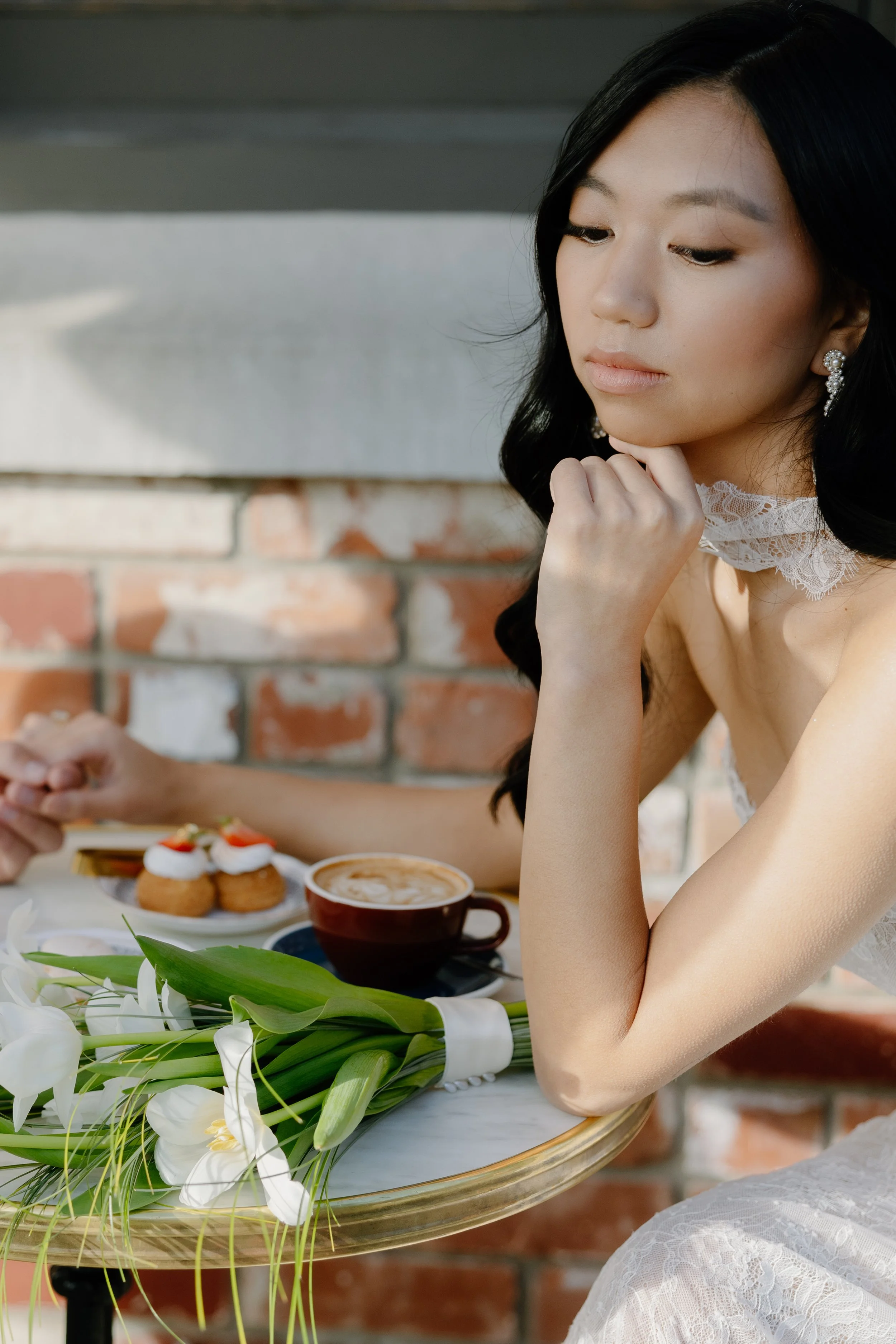 A woman with black hair and earrings seated at a table with a bouquet of white lilies, a coffee cup, and a plate of desserts; brick wall background.