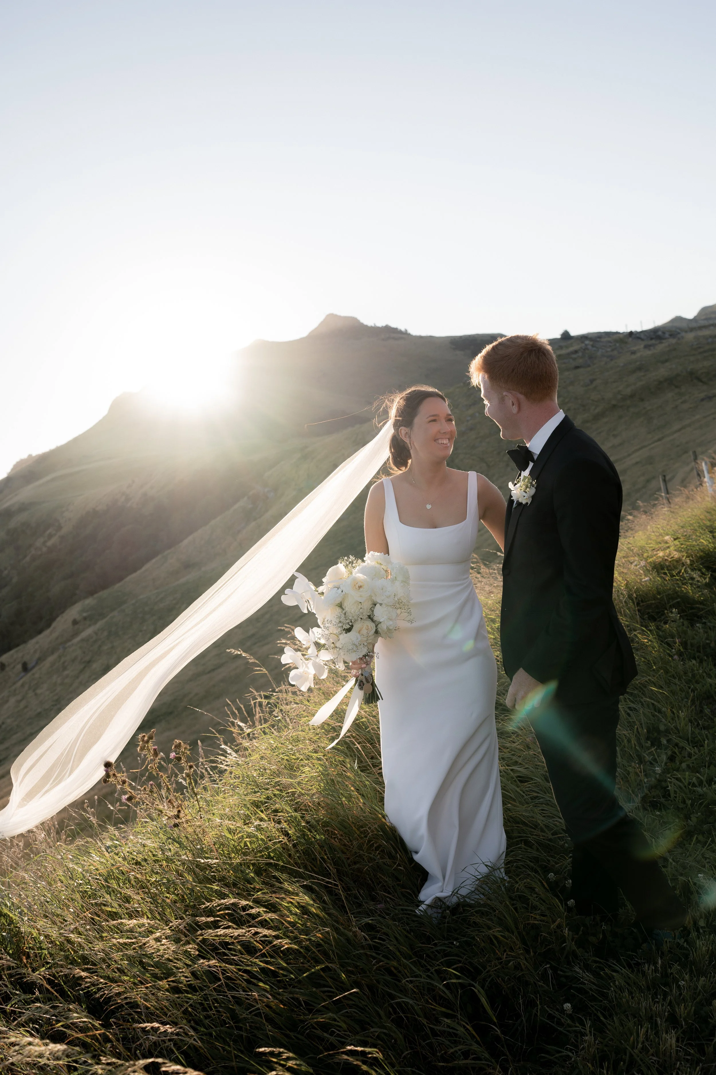 A bride and groom standing outdoors on a grassy hillside at sunset, facing each other and smiling, with mountains in the background.