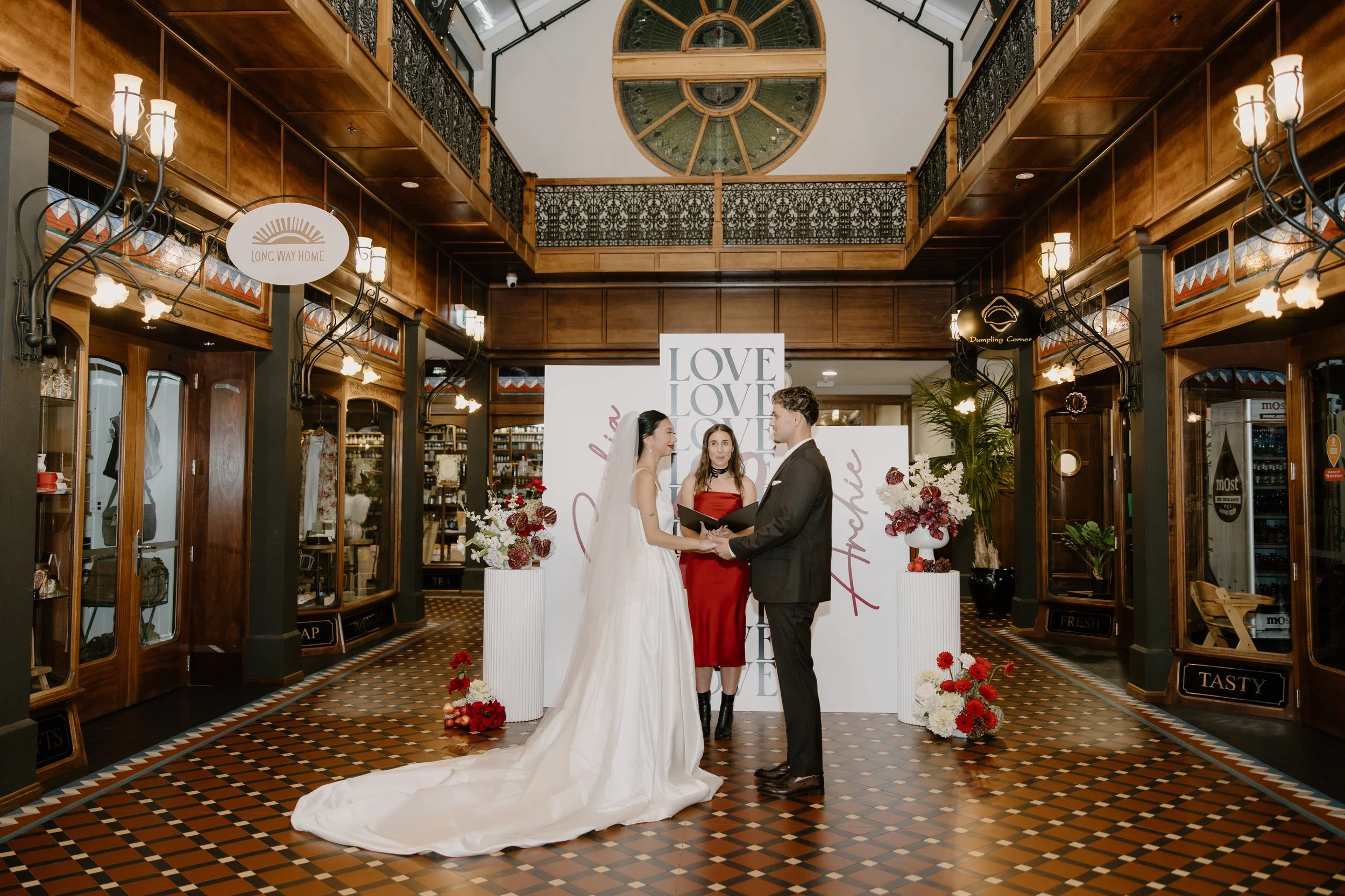 A wedding ceremony inside a historic building with wood-paneled walls and a stained glass ceiling. A bride and groom hold hands and face each other, with a woman standing between them. The area is decorated with flowers and signs reading 'LOVE' and '
