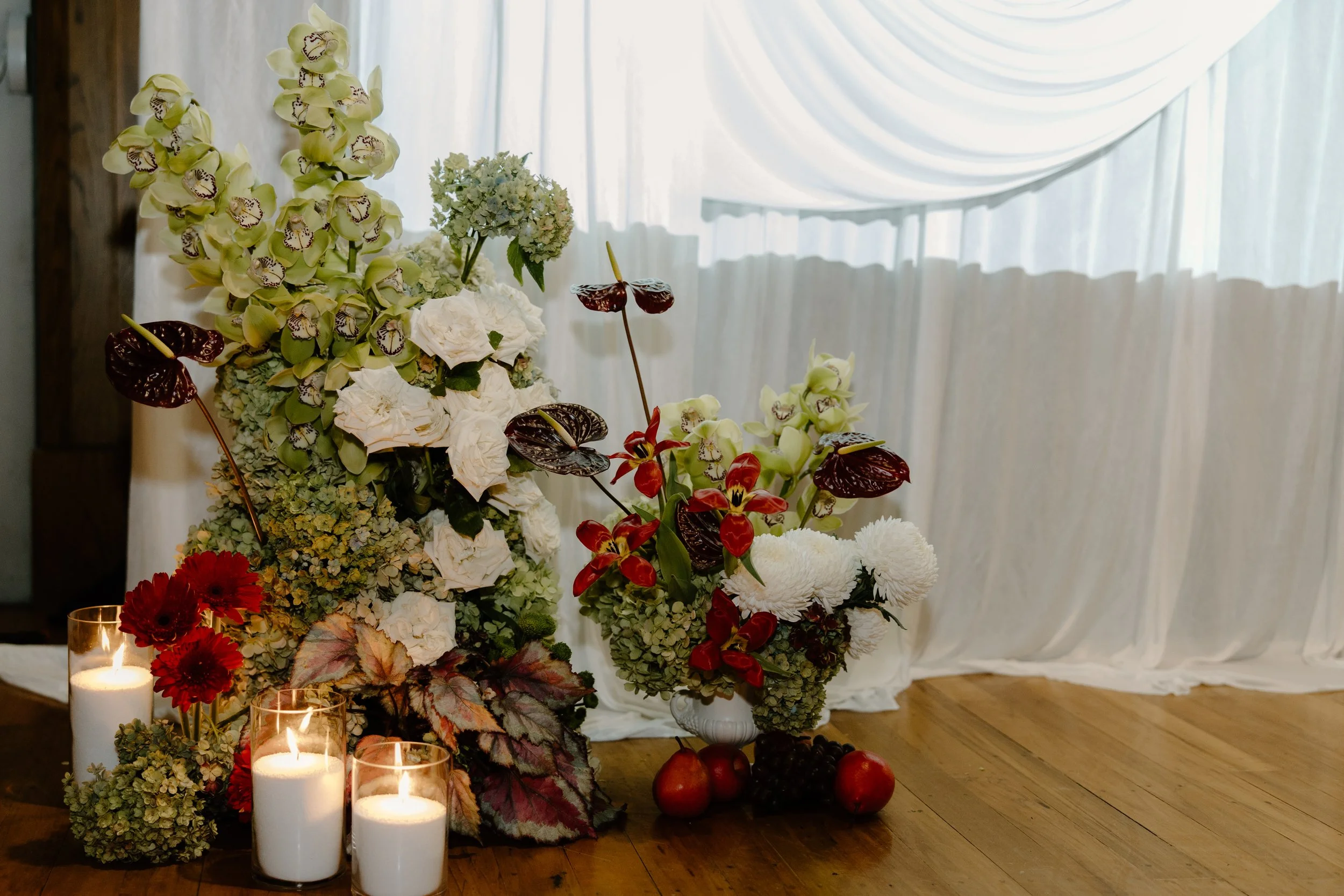 A floral arrangement with white and green flowers, red flowers, and candles in glass holders on a wooden floor in front of white curtains.