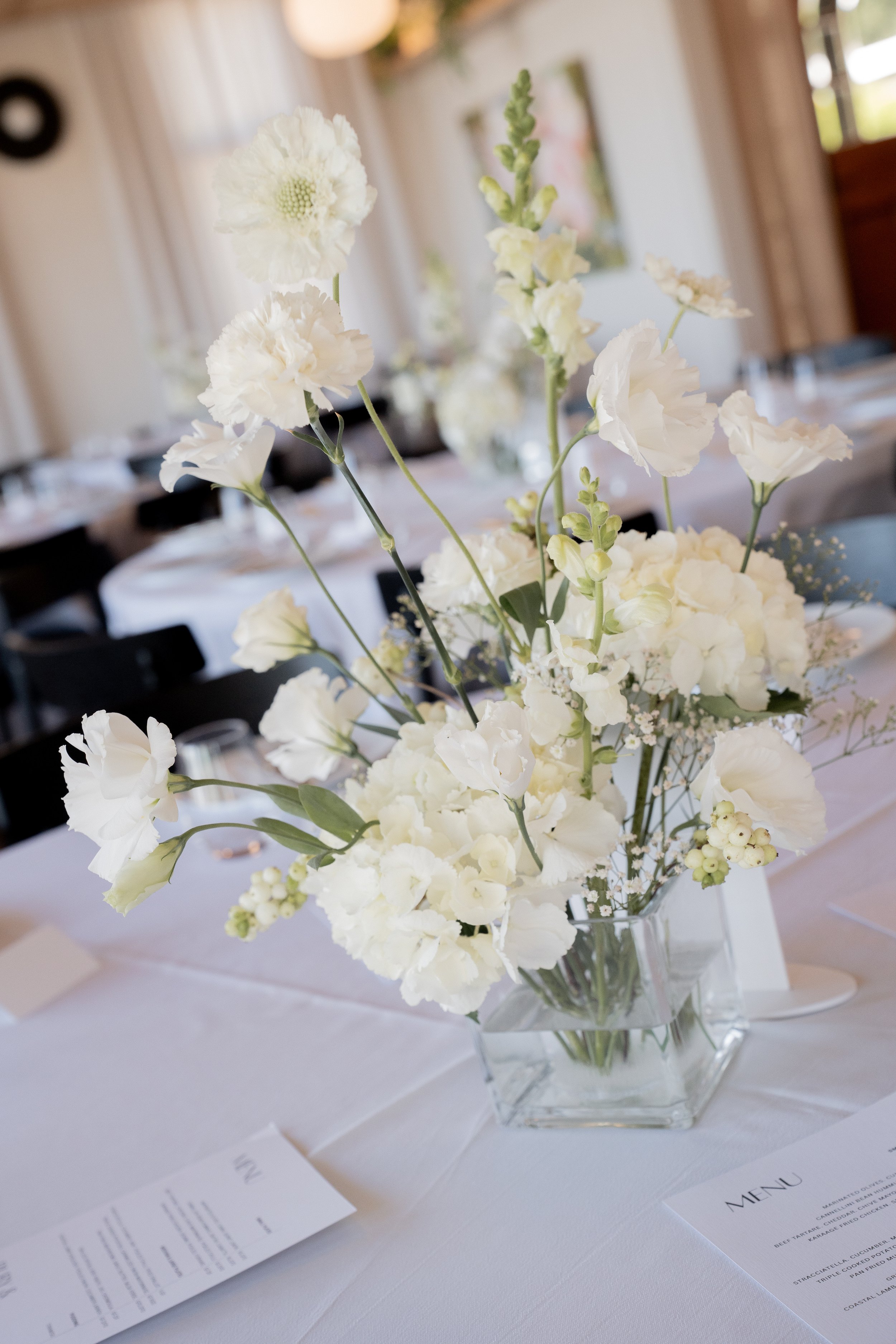 White floral centerpiece with roses and hydrangeas on a dining table with menus for a wedding or event.