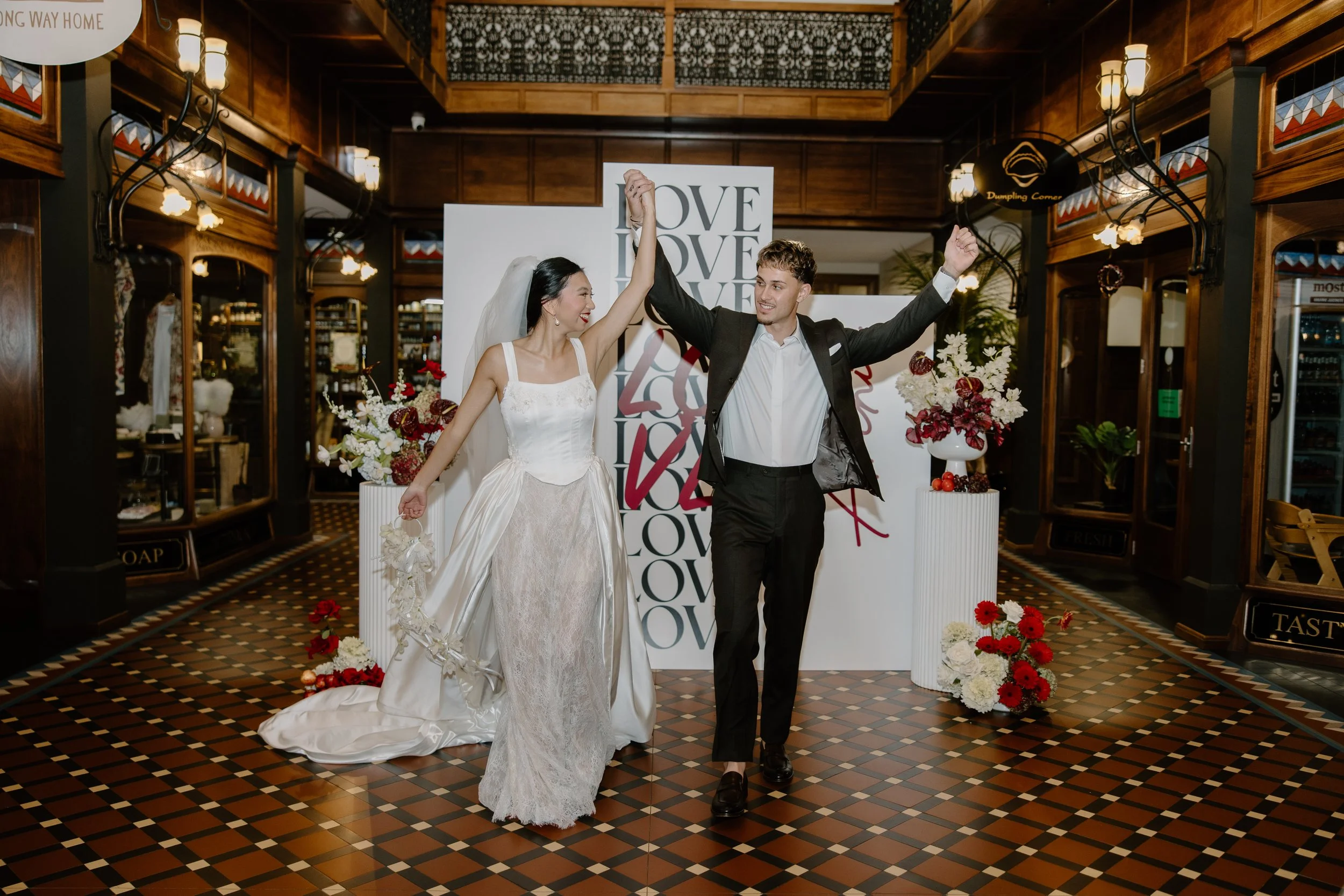 A bride and groom in wedding attire raising their hands together in celebration inside a warmly lit, wood-paneled venue decorated with flowers and a large backdrop with the word 'LOVE' repeated.