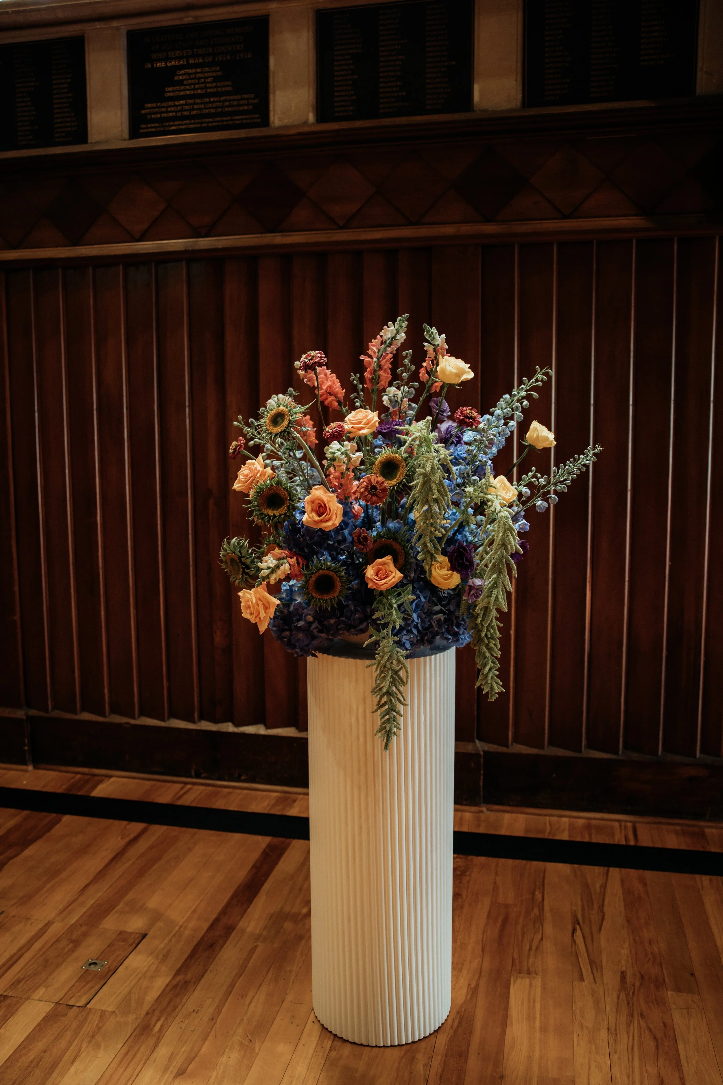 A tall white vase with vertical ridges holds a colorful arrangement of flowers including sunflowers, roses, and other blooms, positioned in front of a dark wooden wall and a plaque in the background.